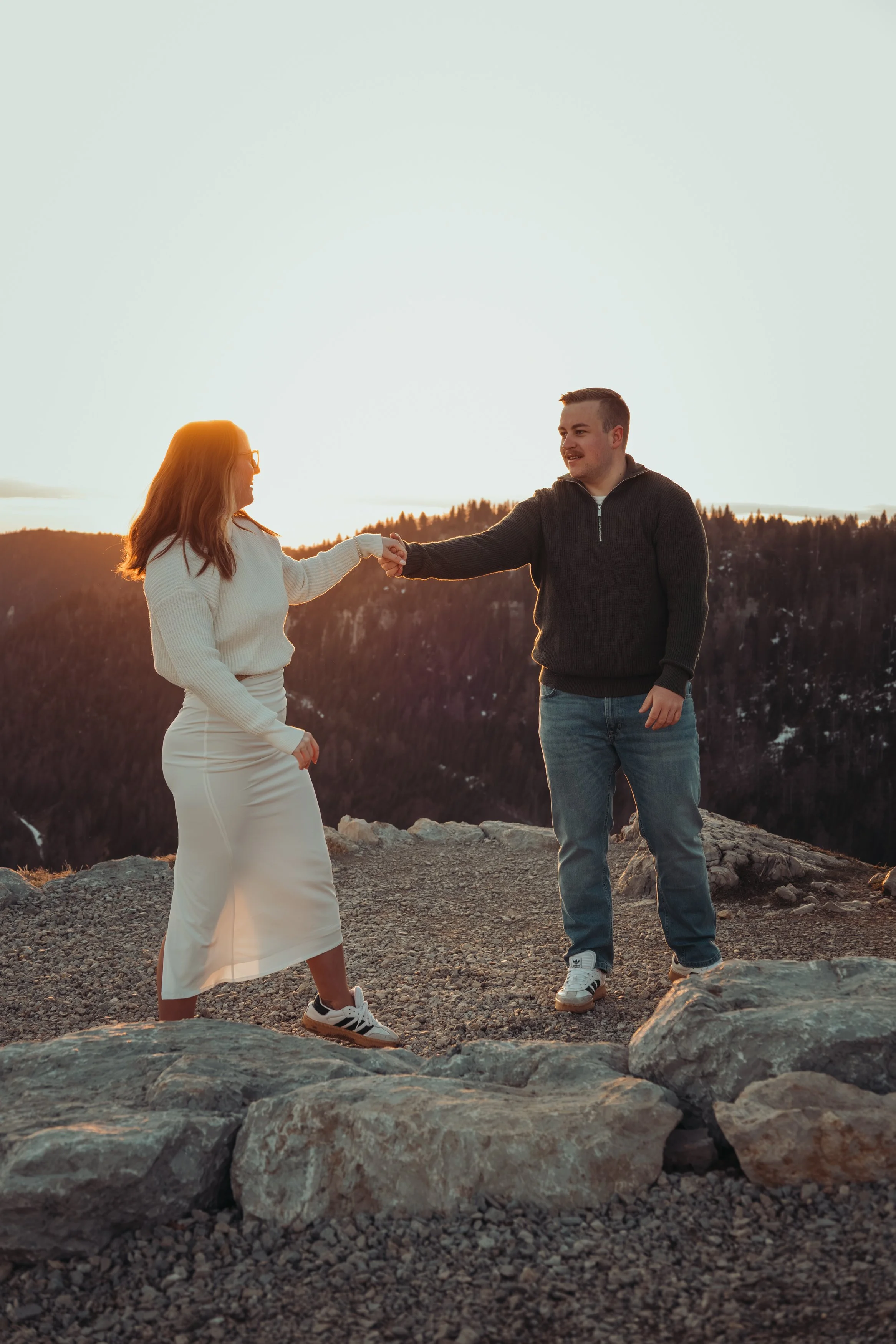 Un homme et une femme échangent une poignée de main en haut d'une montagne lors du coucher du soleil, avec un arrière-plan de forêt et de ciel lumineux.