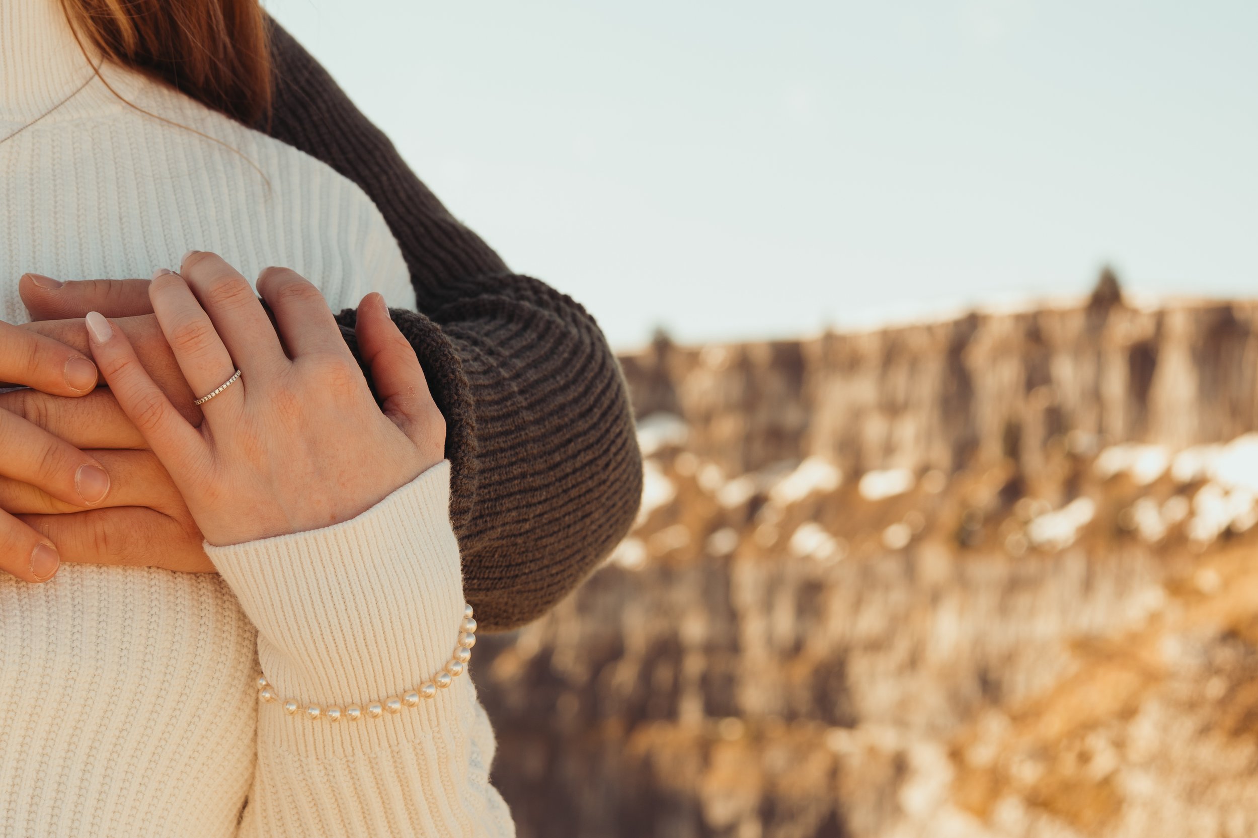 Deux personnes dont les bras sont croisées, une portant un pull blanc et une autre un pull marron, dans un paysage extérieur avec un ciel clair et un mur en pierre en arrière-plan.