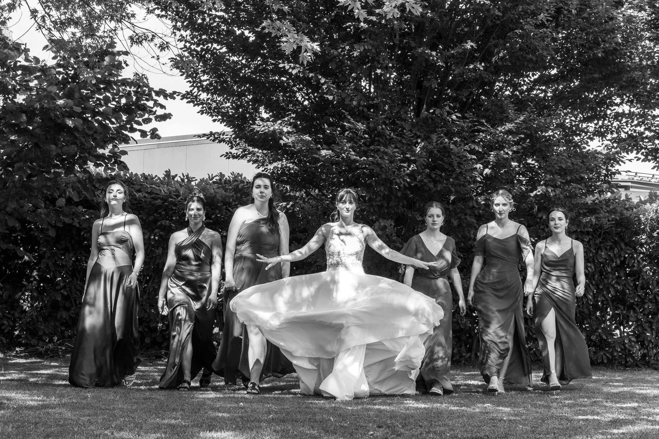 Groupe de sept femmes en robes élégantes posant en extérieur sous un grand arbre, en noir et blanc.