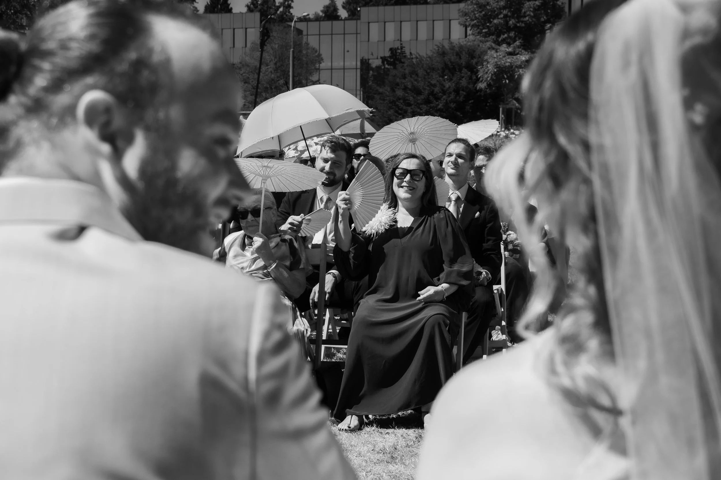 Groupe de personnes assises lors d'une cérémonie en plein air, utilisant des parapluies pour se protéger du soleil, avec des bâtiments modernes en arrière-plan.