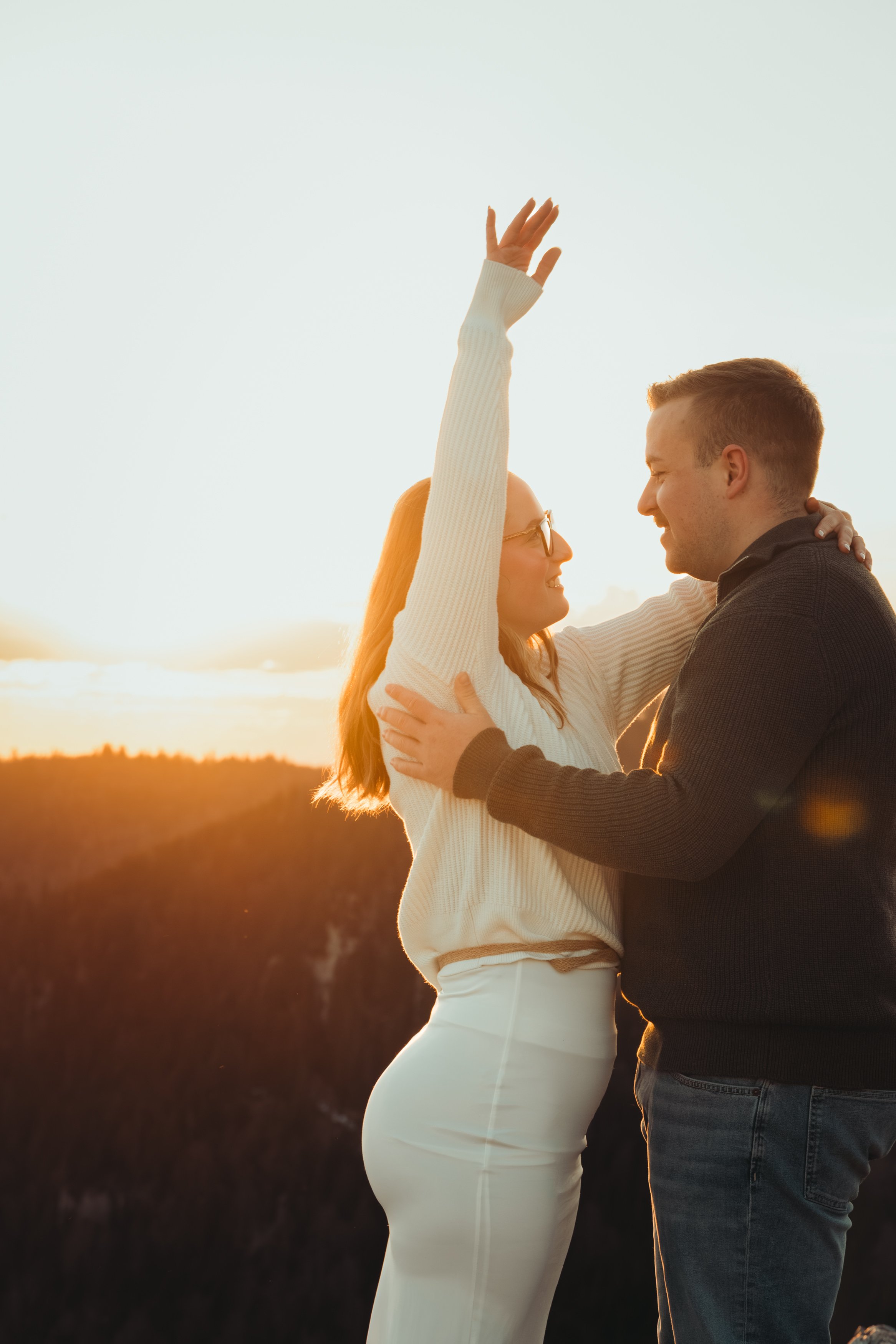 Un couple s'embrasse lors du coucher du soleil, la femme en robe blanche et l'homme en pull noir, dans un paysage naturel.