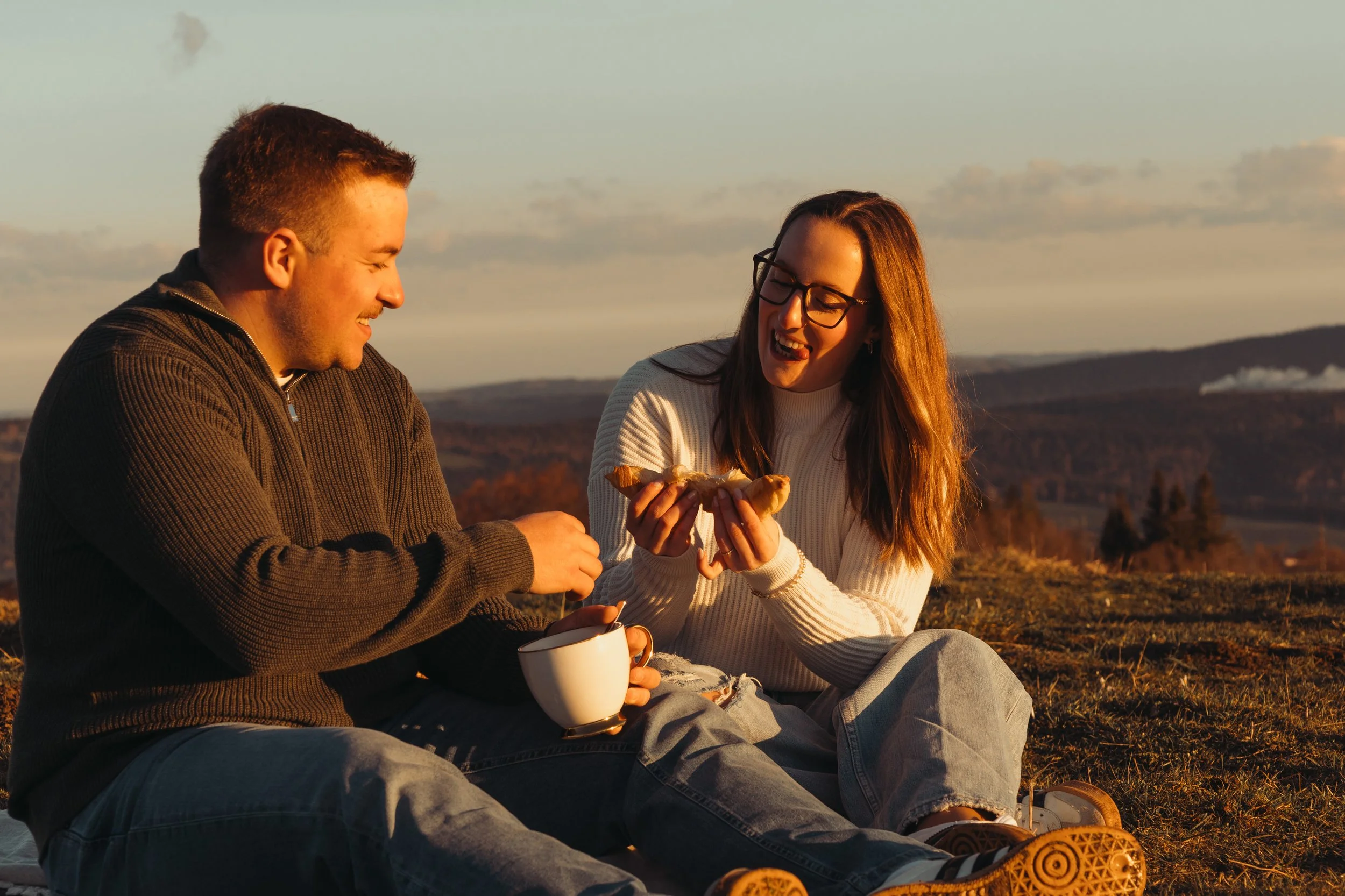 Deux jeunes adultes assis à l'extérieur, partageant un moment joyeux lors d'un coucher de soleil sur un paysage rural ou montagneux. La femme tient un croissant et rit, tandis que l'homme lui donne quelque chose, tous deux souriants.