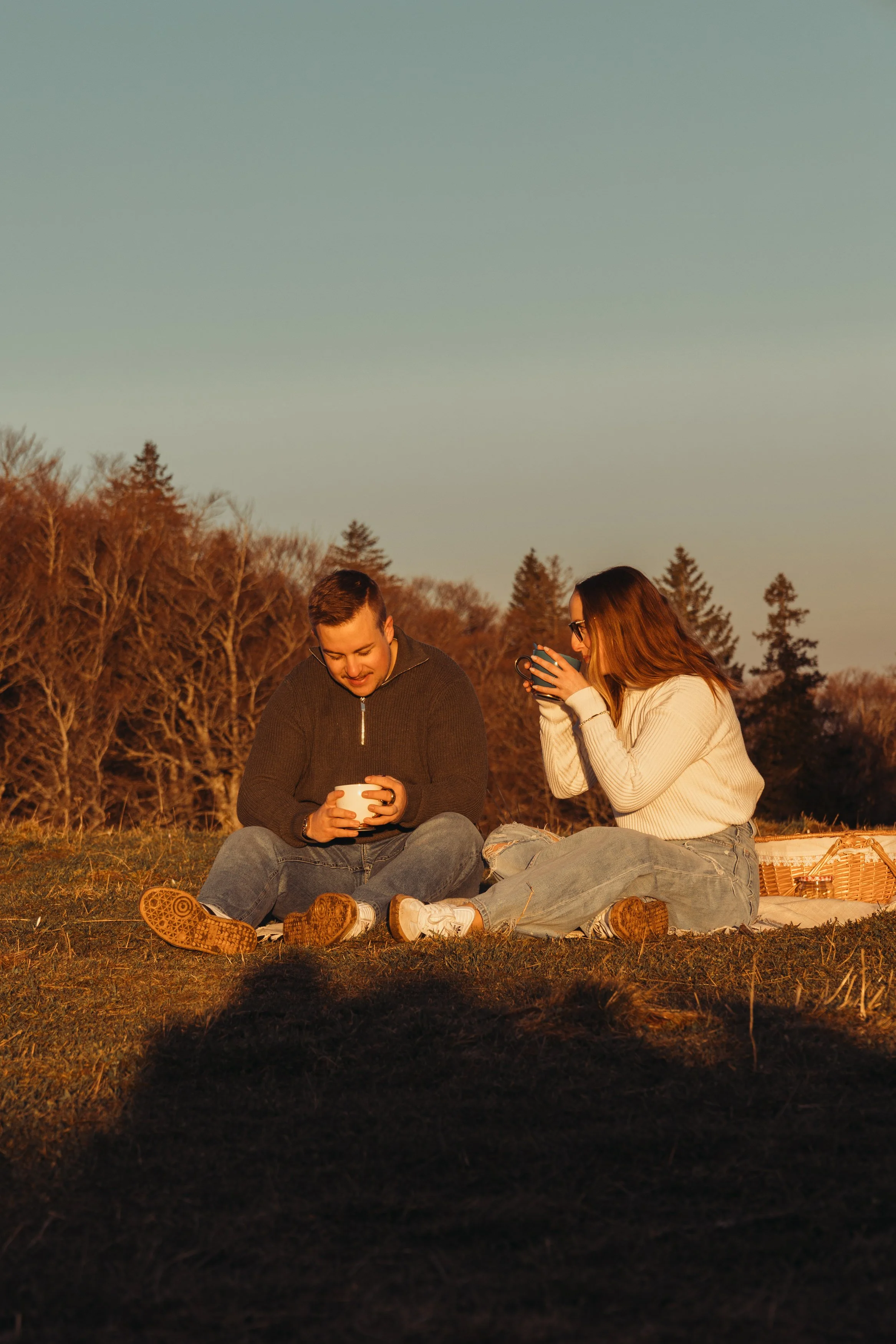 Deux jeunes femmes assises sur l'herbe lors d'un pique-nique en forêt, regardant leurs téléphones, avec un panier à côté, au moment du coucher de soleil.