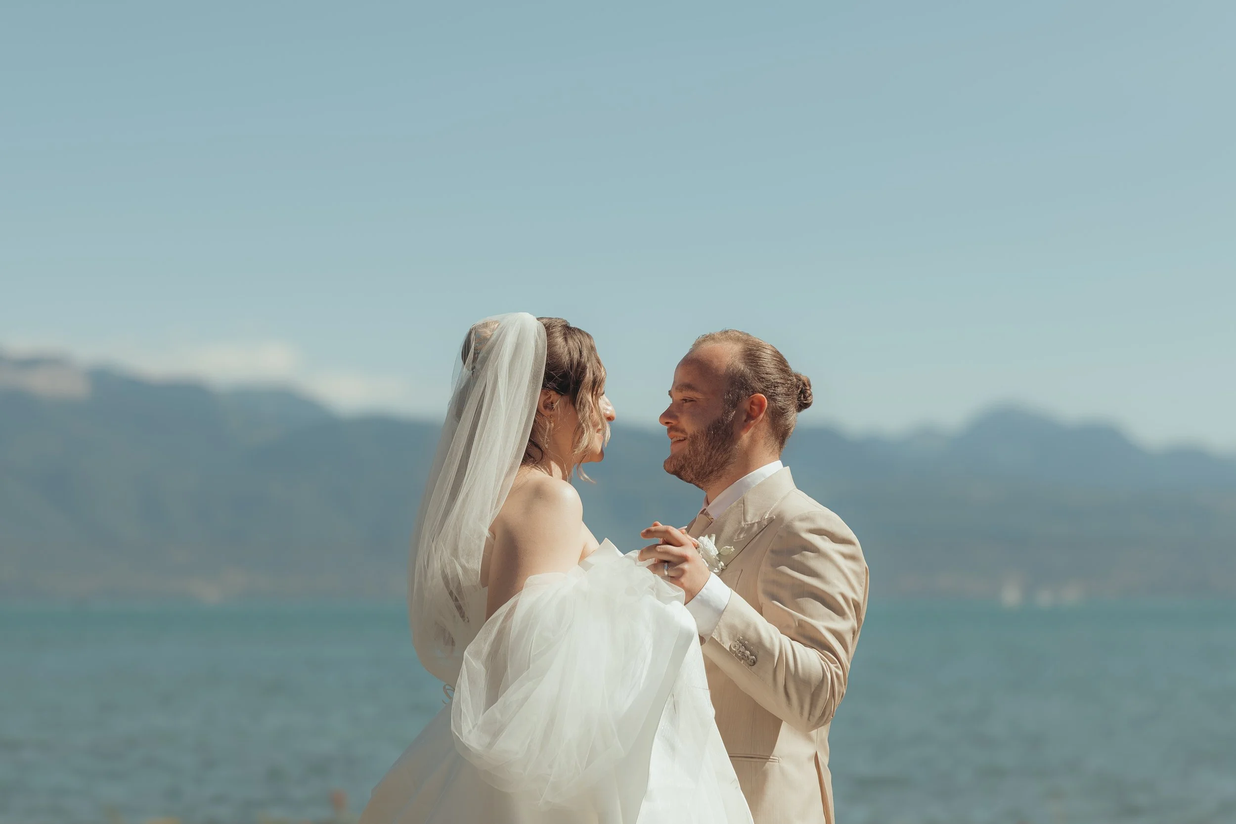 Un couple marié, lui en costume beige et elle en robe de mariée blanche, se regardent tendrement sur une plage avec la mer en arrière-plan et des montagnes lointaines sous un ciel bleu clair.