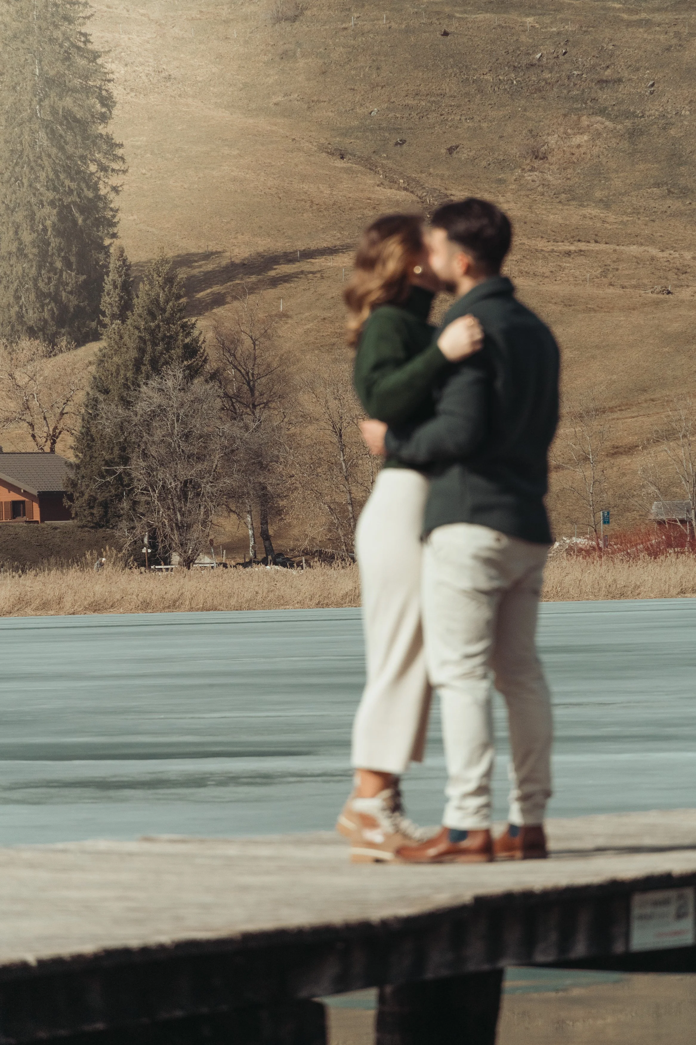 Un couple partage un moment intime en se tenant par la taille, regardant chacun dans les yeux, dans un paysage champêtre avec des arbres et une maison en arrière-plan.