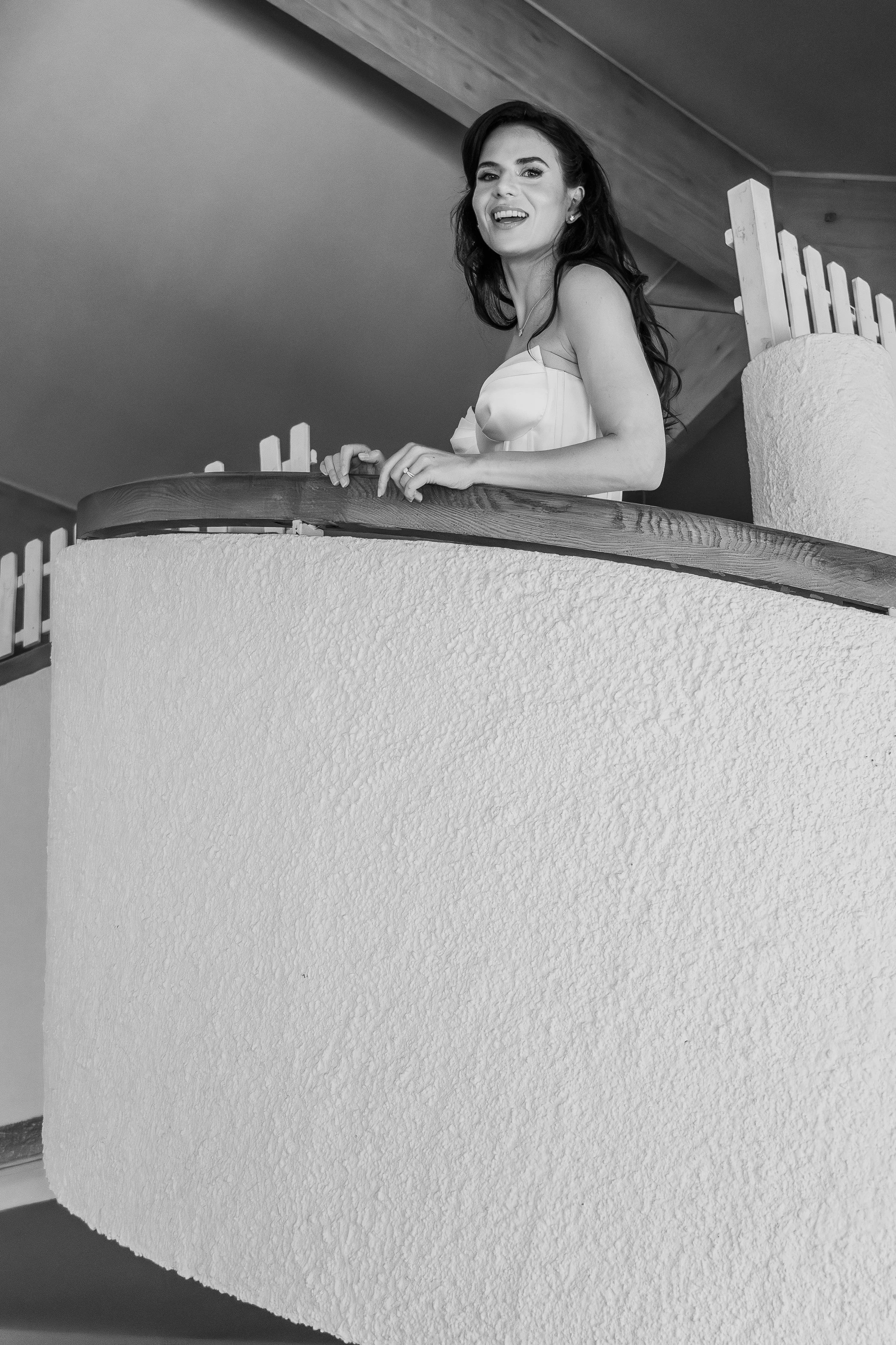 Une femme en robe blanche sourit en regardant vers l'appareil photo, debout sur un balcon avec une rambarde en bois et une surface en plâtre à l'avant-plan.