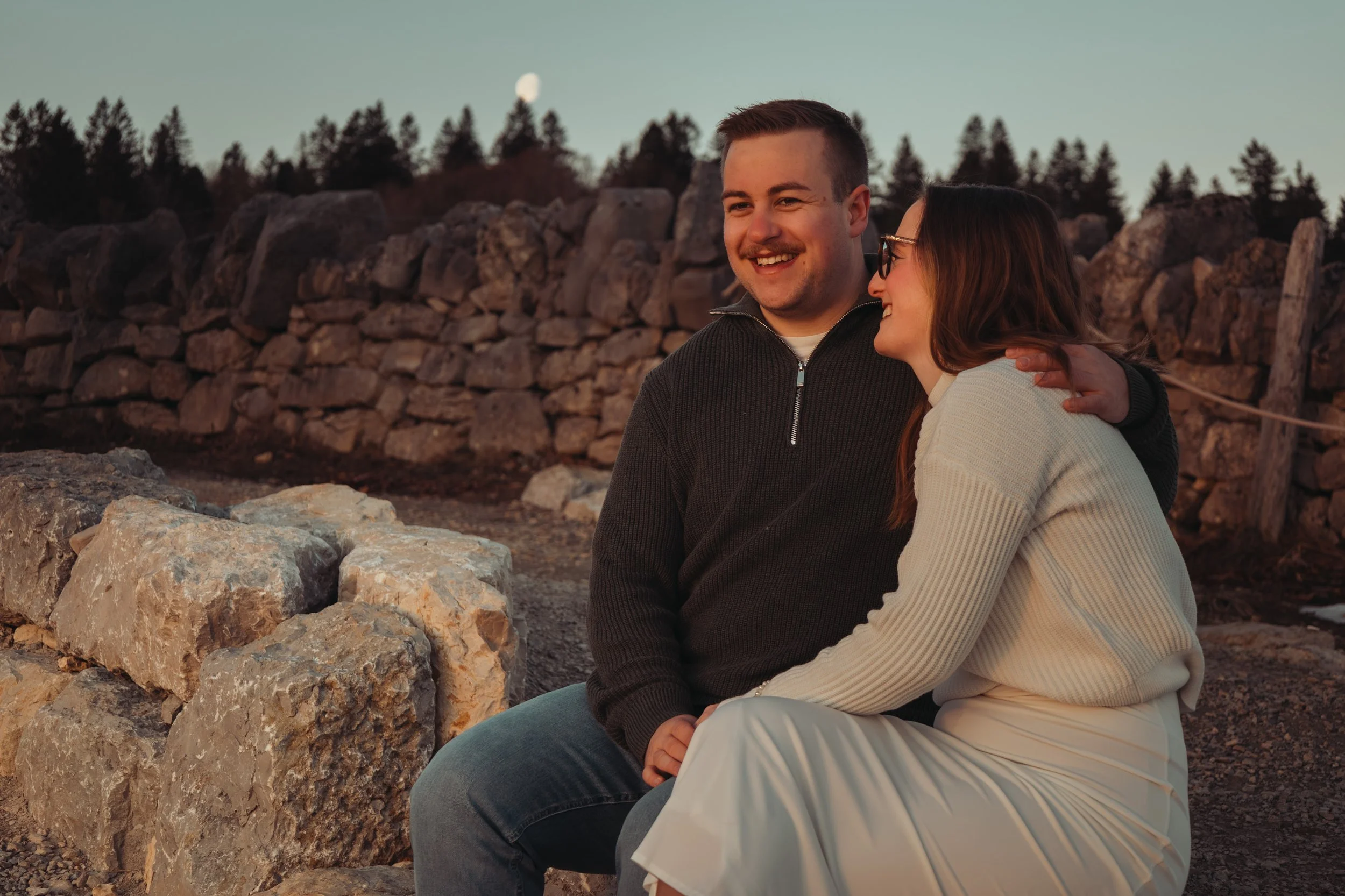 Deux personnes assises sur des rochers près d'un mur en pierre, souriantes, en plein air au crépuscule avec la lune visible dans le ciel.