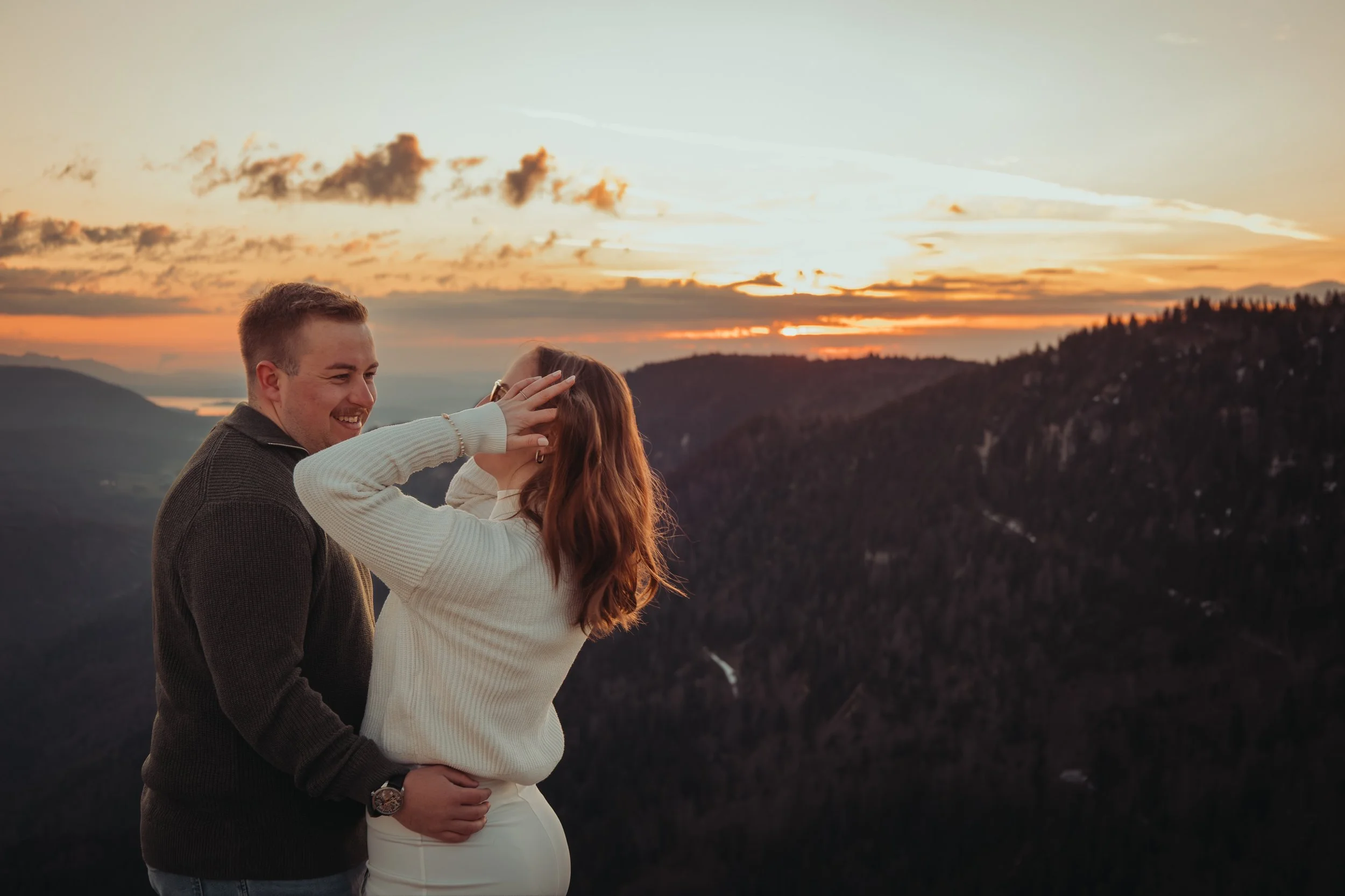 Un couple souriant, en regardant une vue panoramique d'un coucher de soleil avec des montagnes et un lac en arrière-plan.