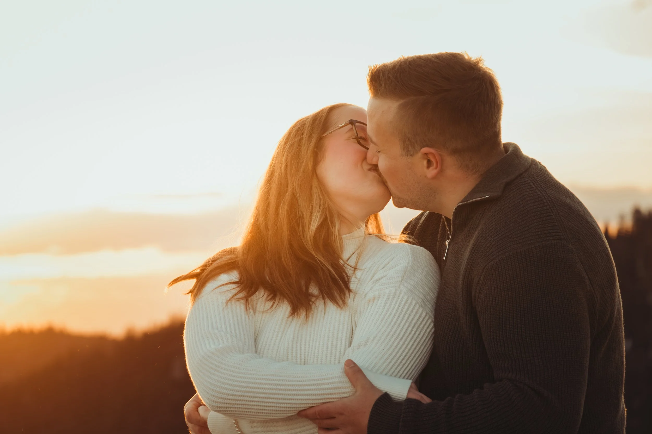 Un couple s'embrasse tendrement lors d'un coucher de soleil à la plage, avec des cheveux roux et bruns, portant des vêtements confortables, dans une ambiance chaleureuse et romantique.