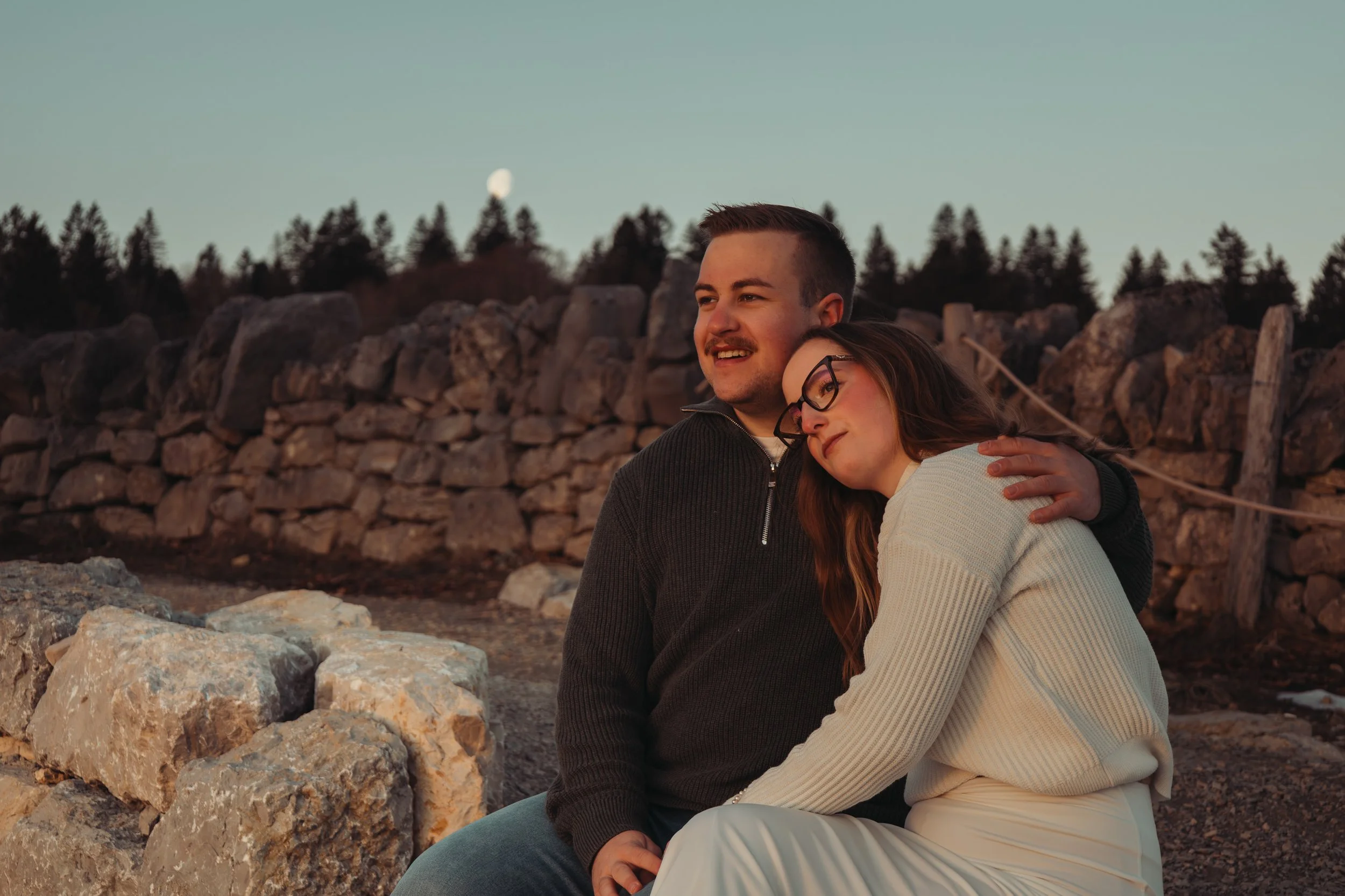 Un couple assis sur des rochers près d'un mur de pierres, profitant du coucher de soleil, avec la lune visible dans le ciel clair.