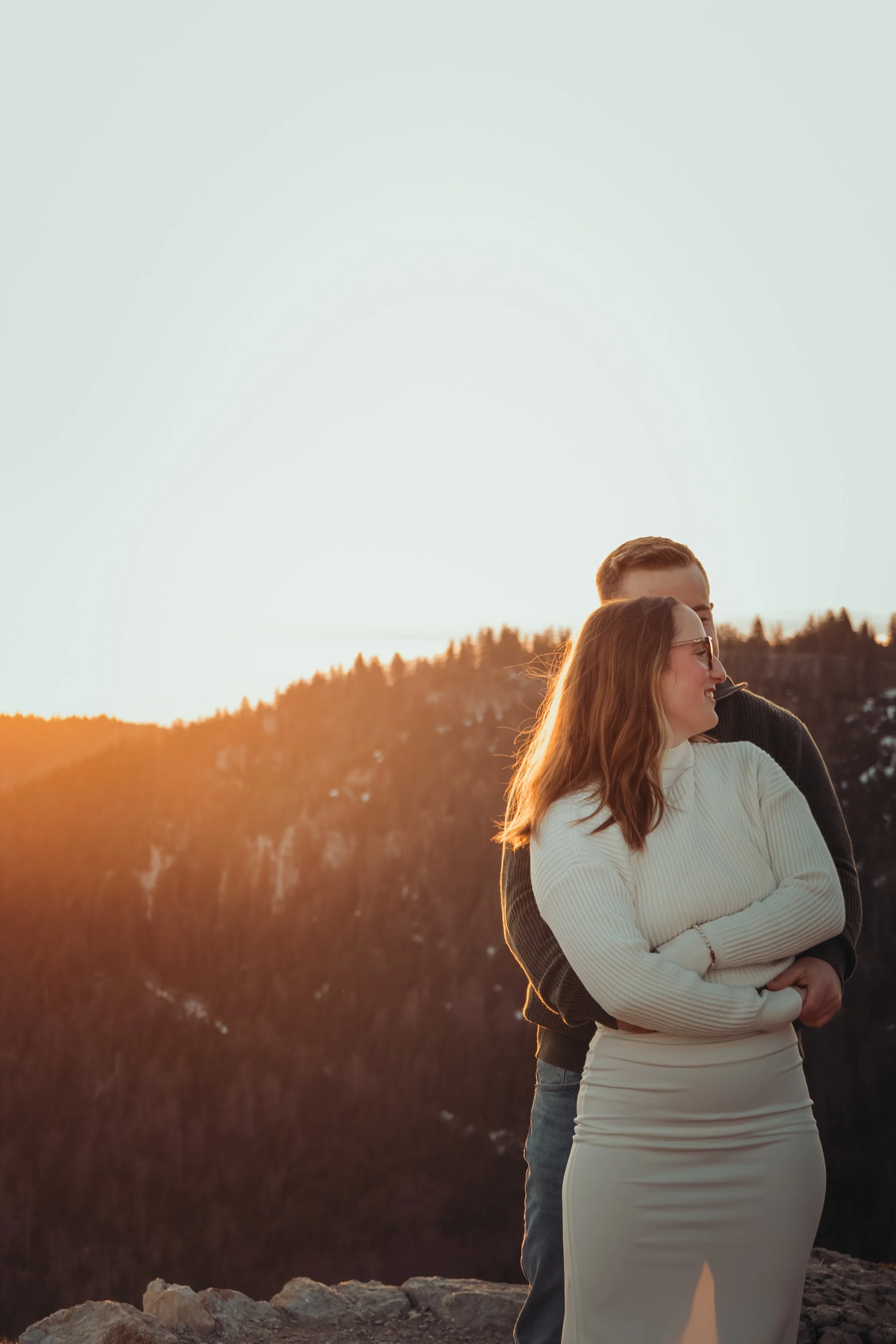 Un couple debout près d'une falaise au coucher du soleil, avec la femme qui porte une robe longue beige et l'homme dans une veste noire, souriant et profitant du paysage.