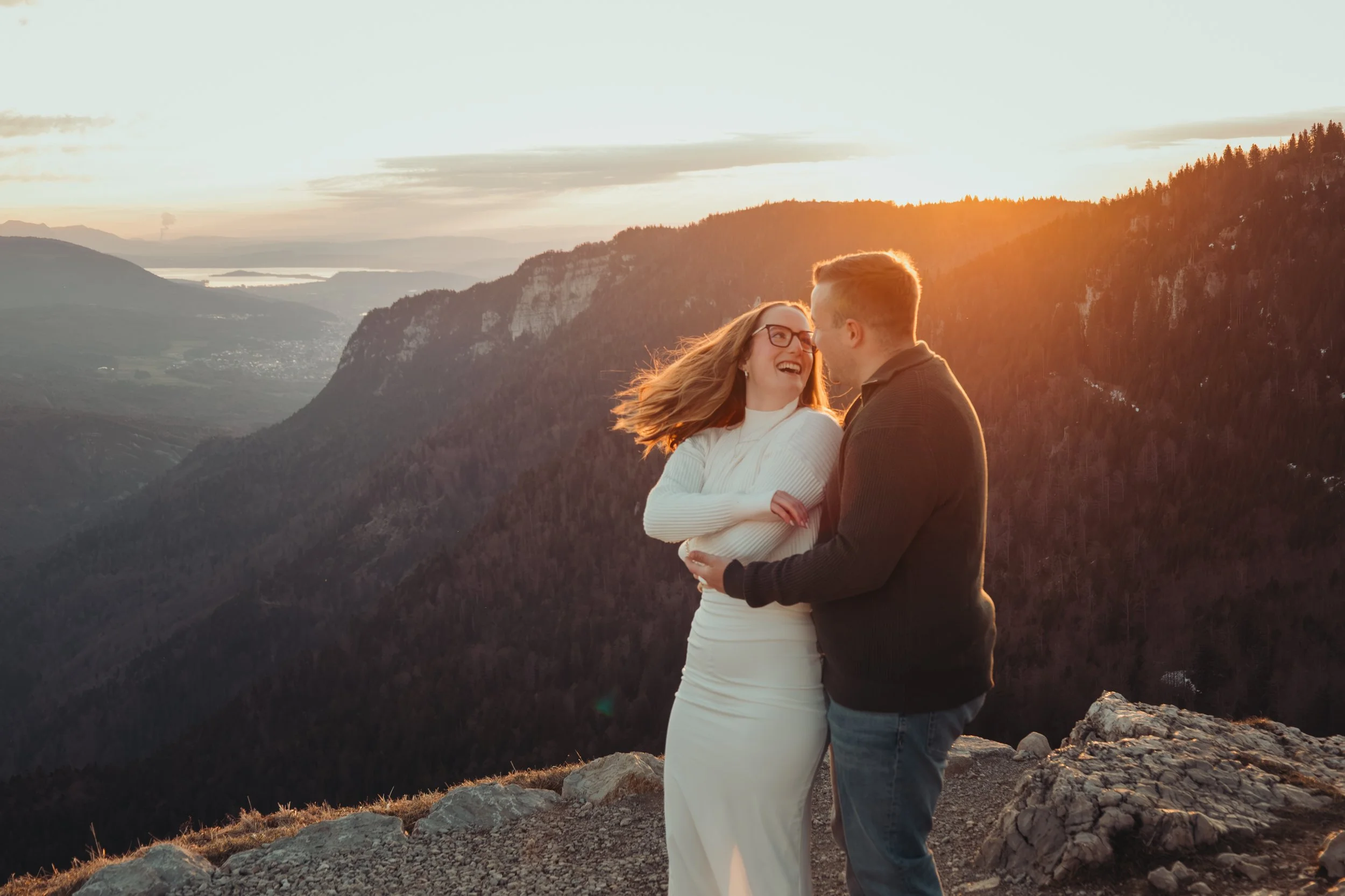 Un couple souriant se tenant la main et se faisant un câlin lors du coucher du soleil, avec une vue sur des montagnes et une vallée en arrière-plan.