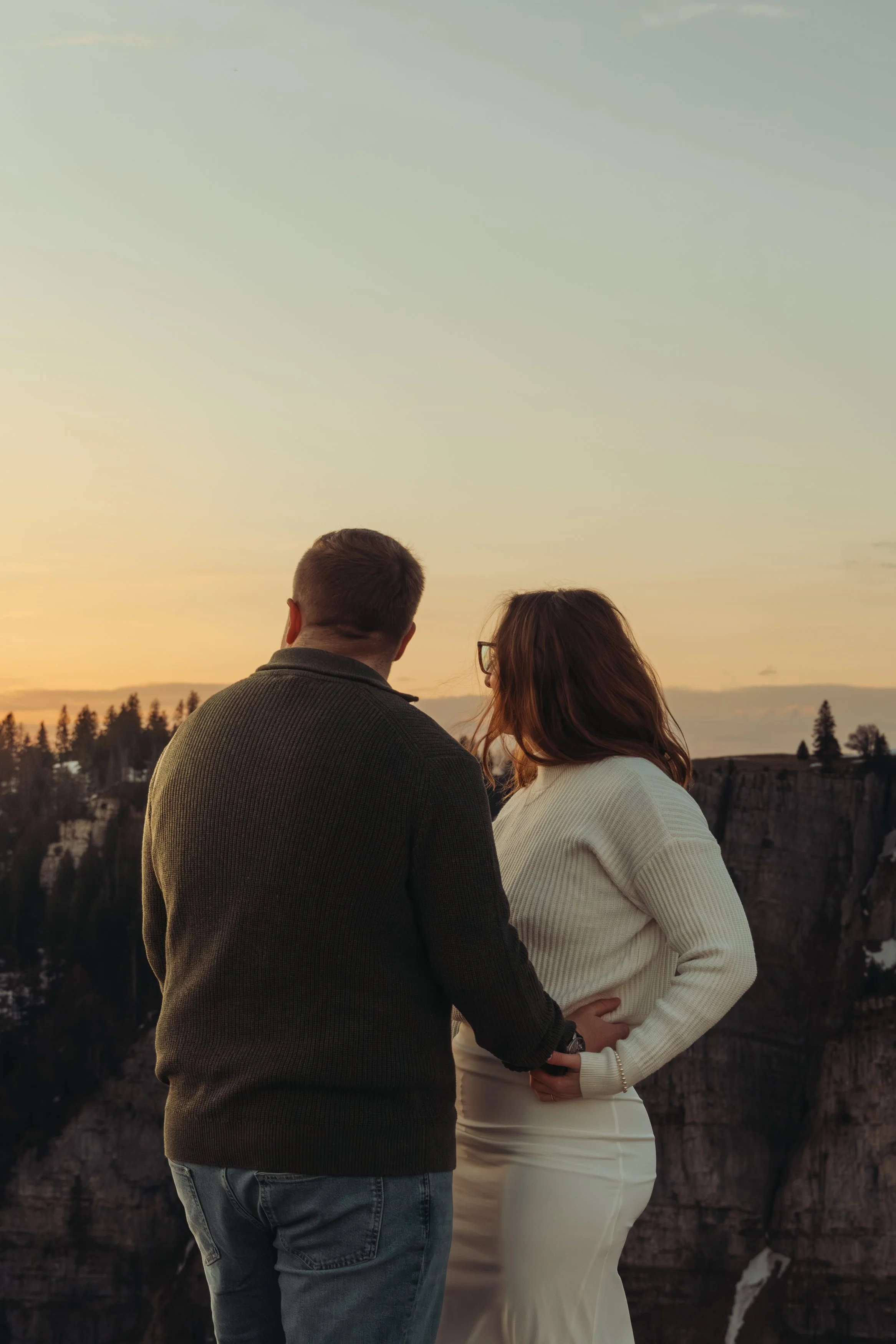 Un couple se tient près d'une falaise au coucher du soleil, regardant au loin. La femme porte un pull blanc et la robe blanche. L'homme porte un pull gris foncé et un jean. Ils sont vus de dos, avec des arbres et la vallée en arrière-plan.