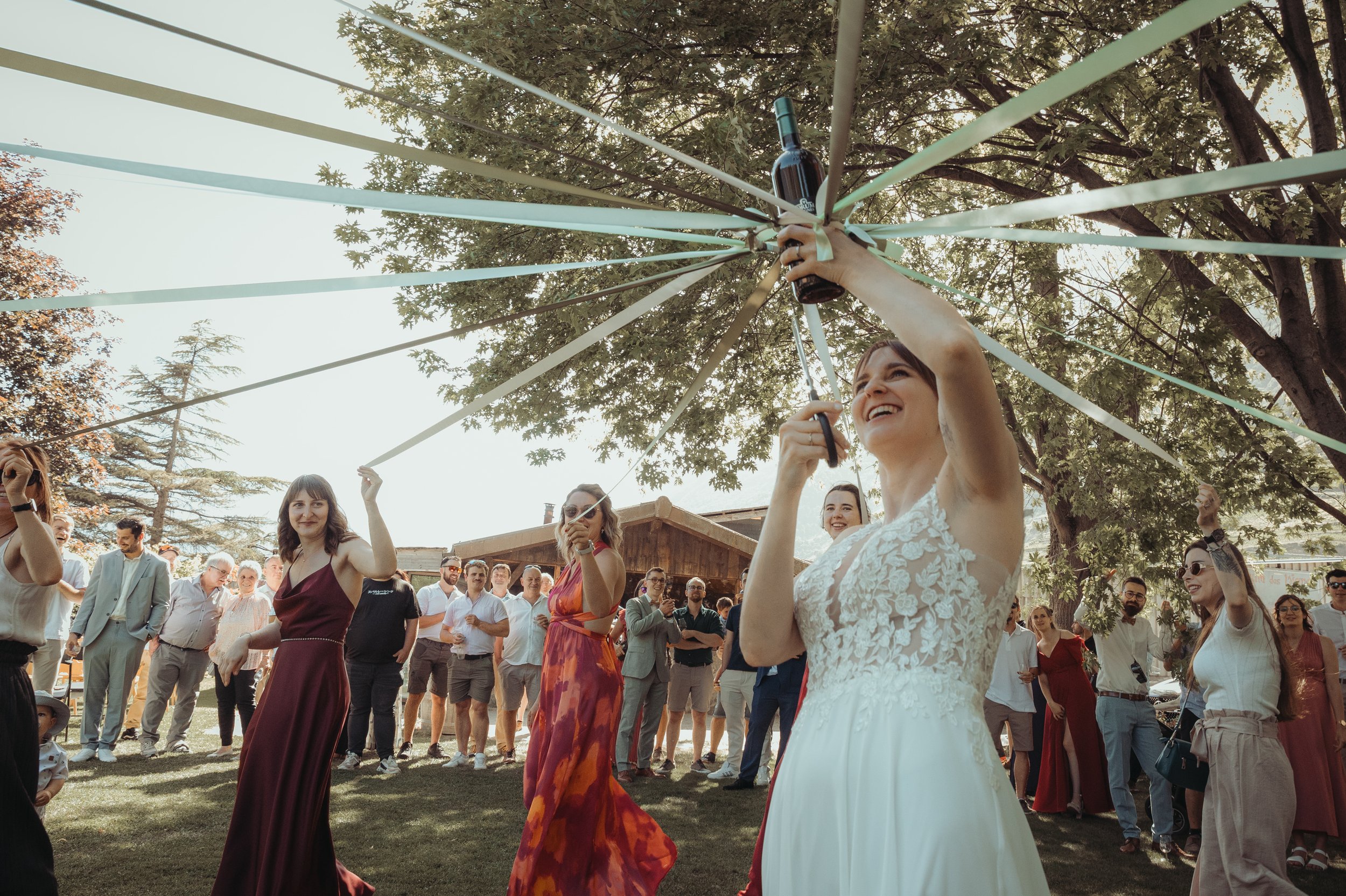 Un groupe de personnes assiste à une célébration en plein air, avec une femme en robe de mariée en avant, tenant une bouteille de vin, sous un parasol blanc. Des arbres et un bâtiment en bois sont visibles en arrière-plan.