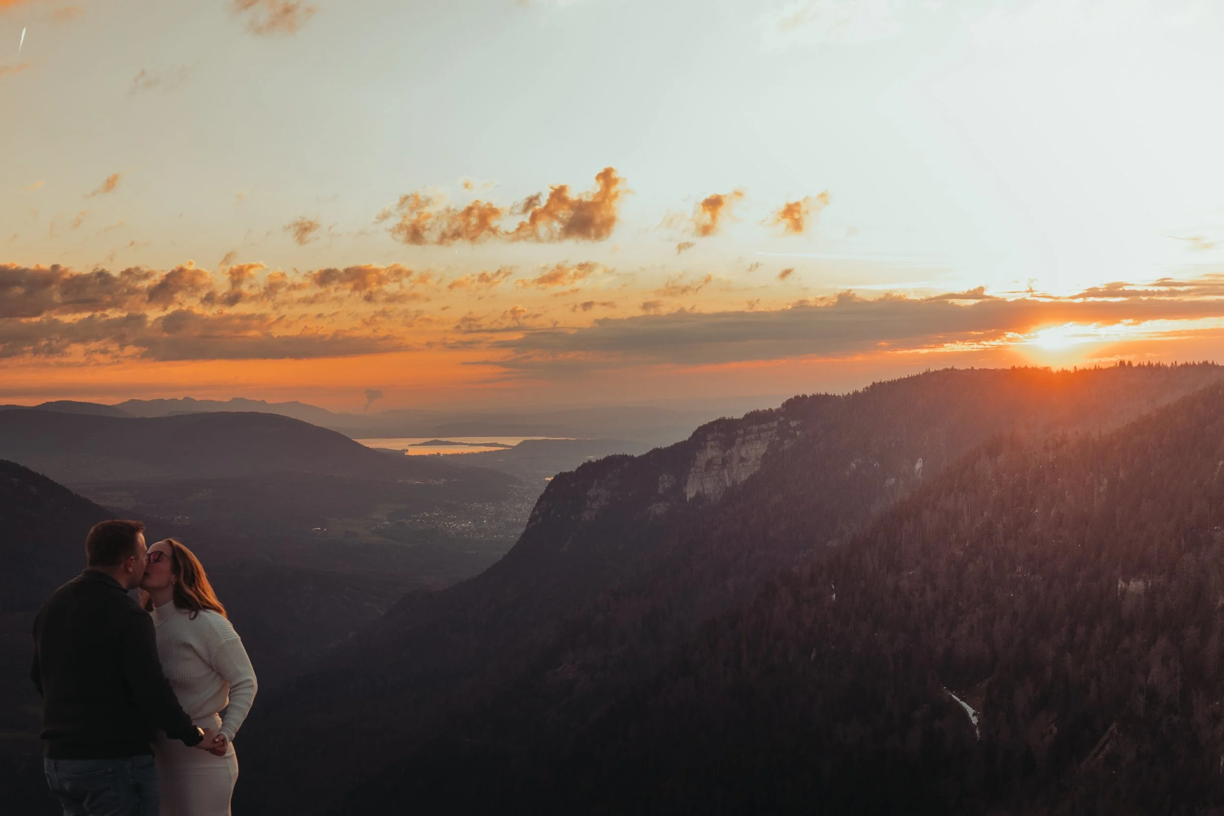 Un couple s'embrasse sur un panorama de montagnes au coucher du soleil, avec un ciel nuageux orange et jaune et une rivière en contrebas.