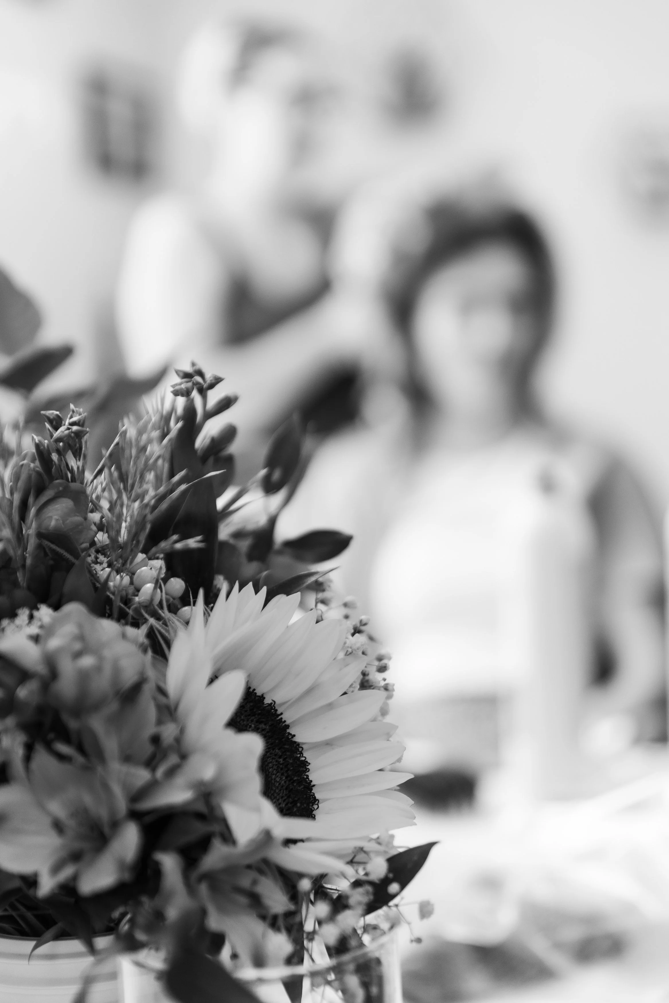 Un bouquet de fleurs, notamment des tournesols, placé dans un vase. En arrière-plan, une personne floue portant des lunettes et un haut à manches longues semble regarder vers le bouquet.