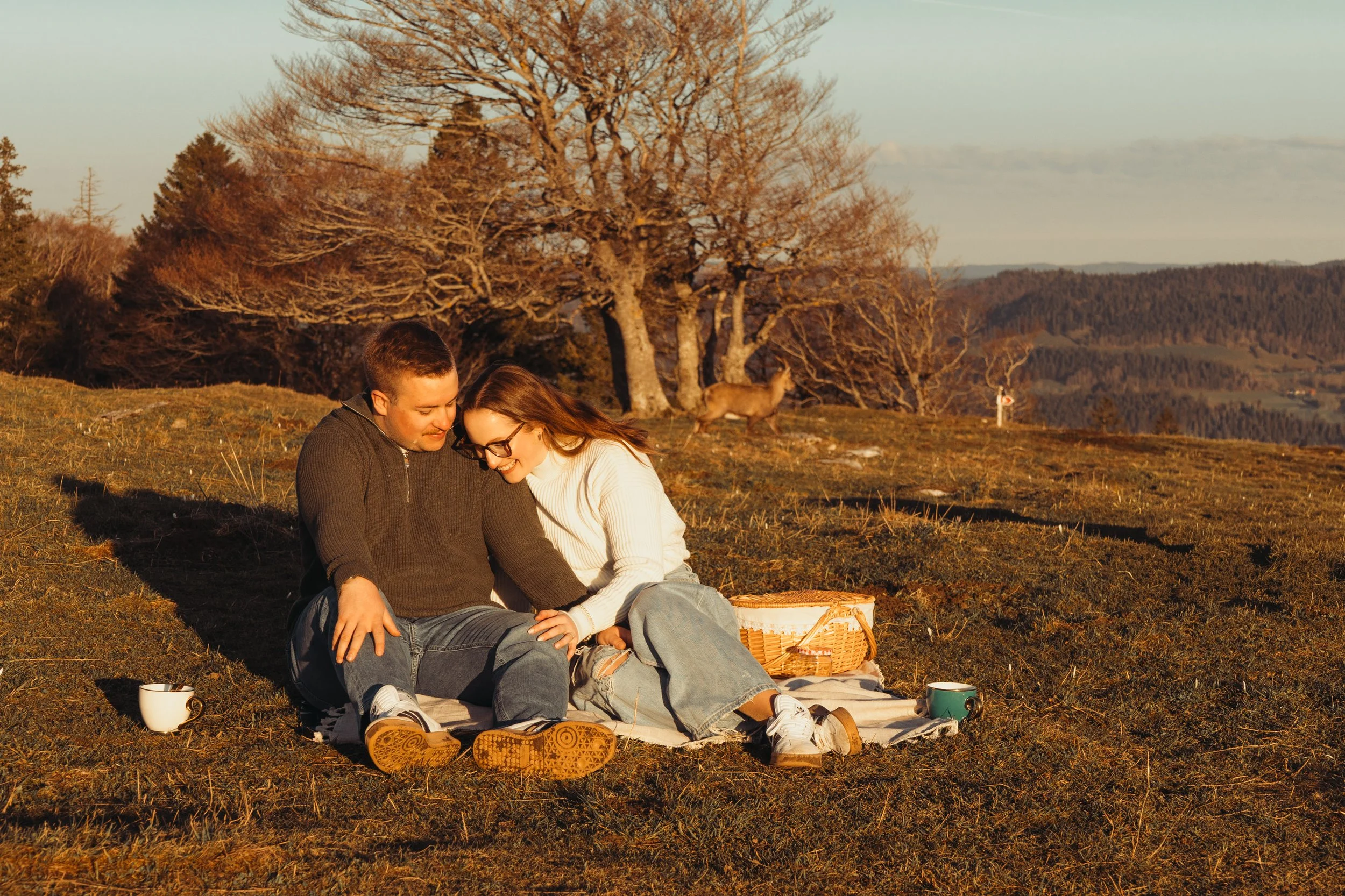 Un couple assis sur une couverture dans un champ en pleine nature, souriant et partageant un moment tendre, avec un cerf en arrière-plan et des arbres dénudés sous une lumière chaude de fin d'après-midi.