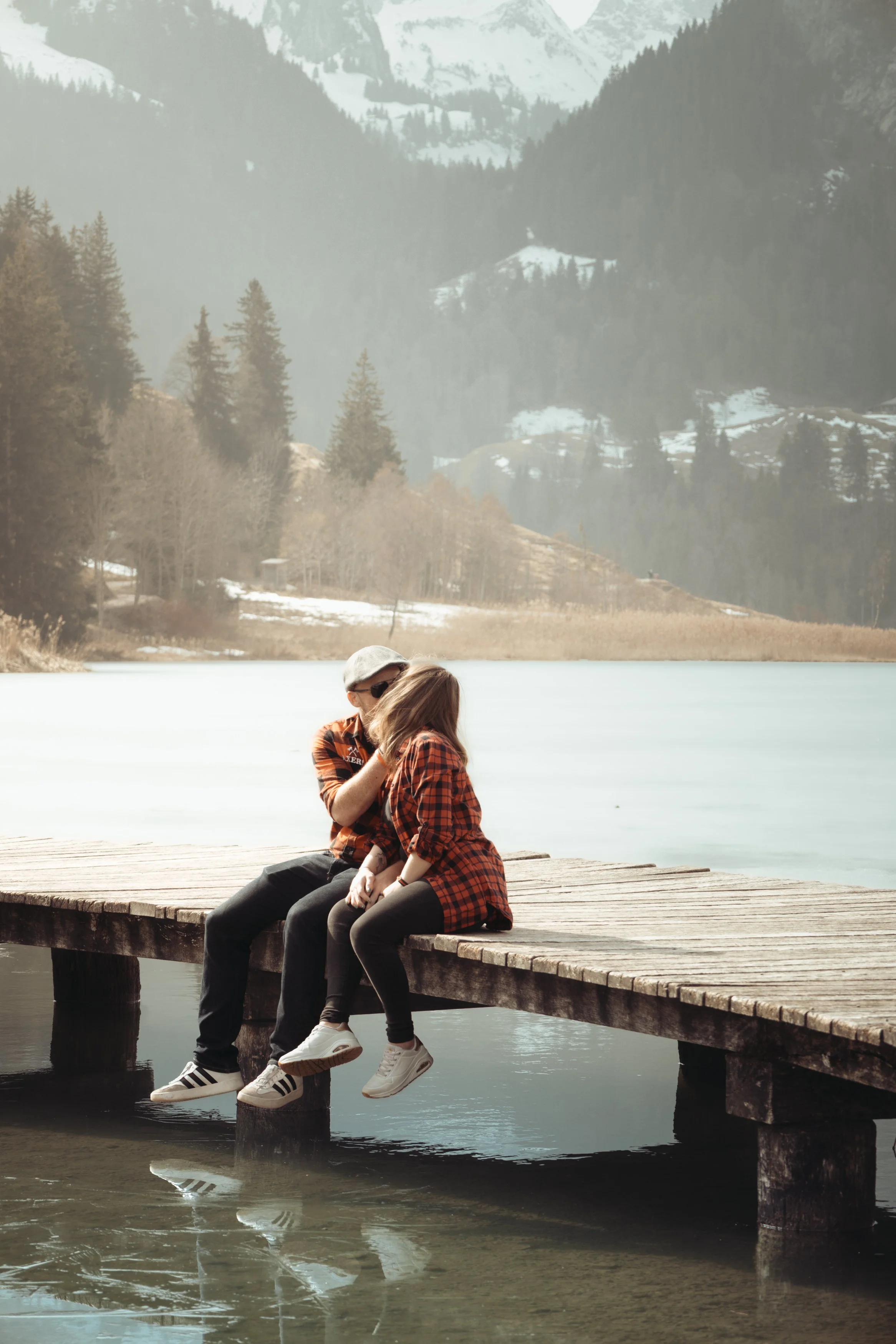 Un couple assis sur une jetée en bois au bord d'un lac, avec une forêt et des montagnes enneigées en arrière-plan.