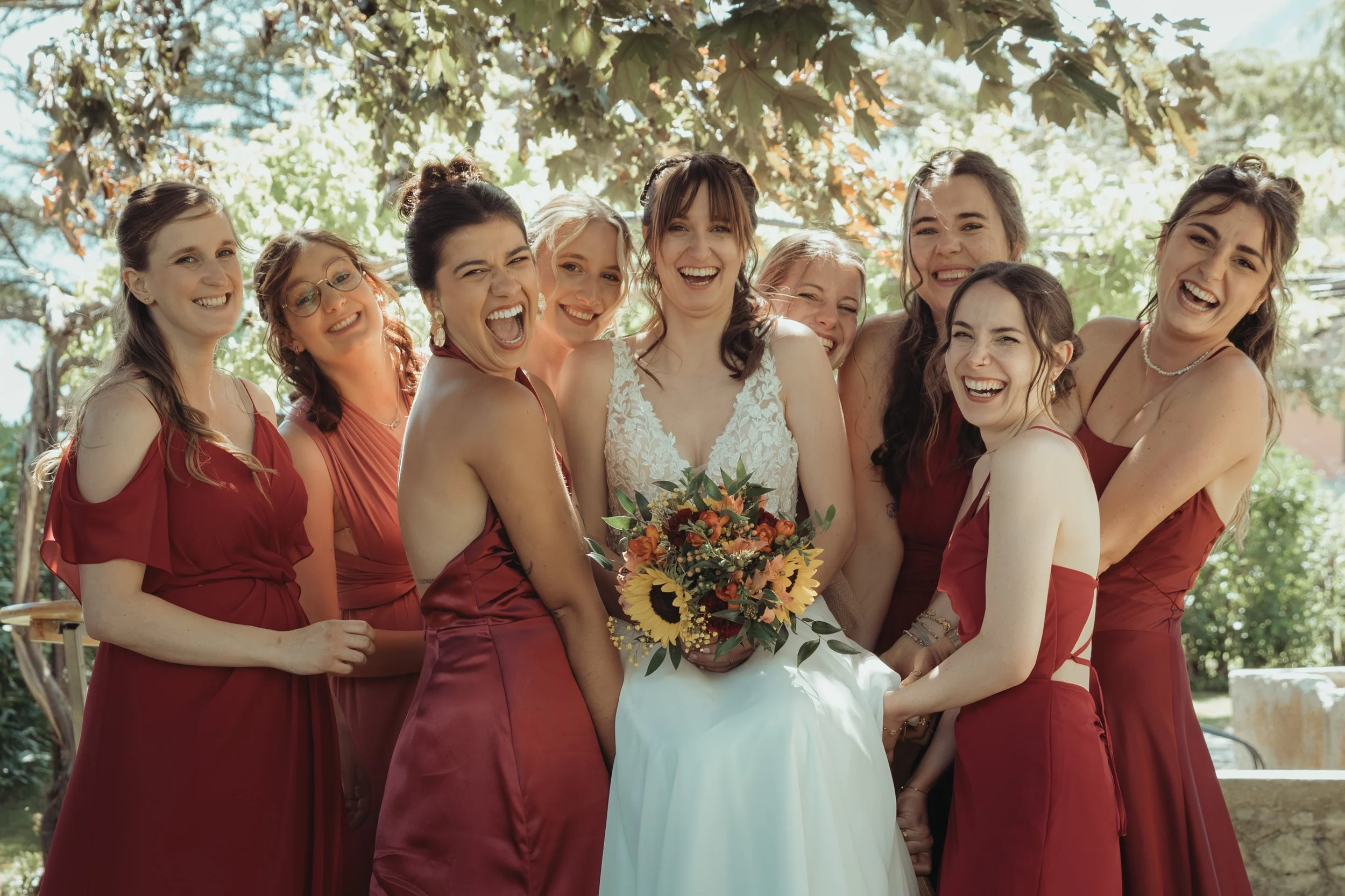 Groupe de femmes souriantes lors d'une célébration en plein air, portant des robes rouges et une robe blanche, avec une femme au centre tenant un bouquet de fleurs