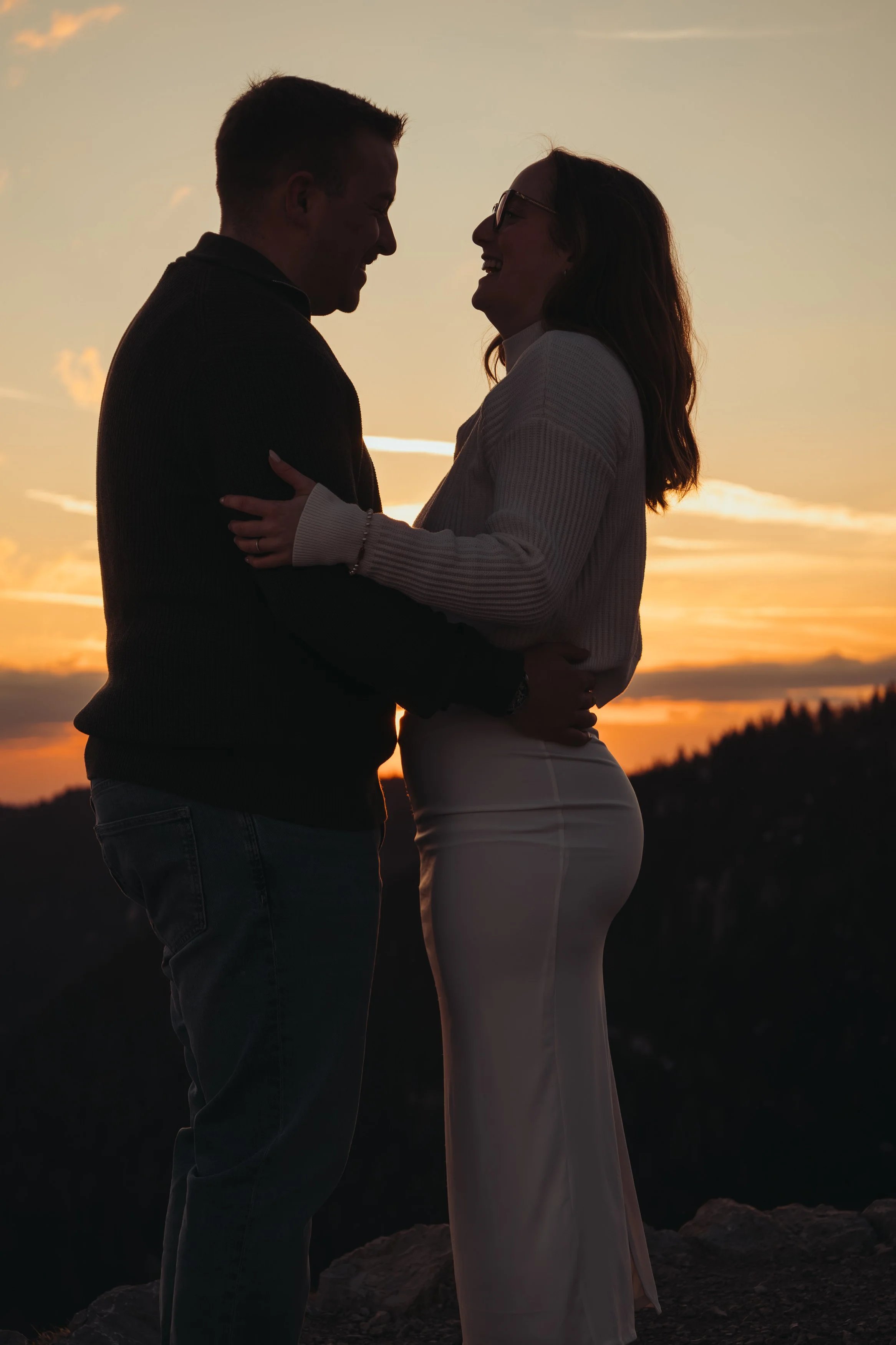 Un couple souriant se regarde tendrement à l'extérieur au coucher du soleil, avec un paysage de collines en arrière-plan.