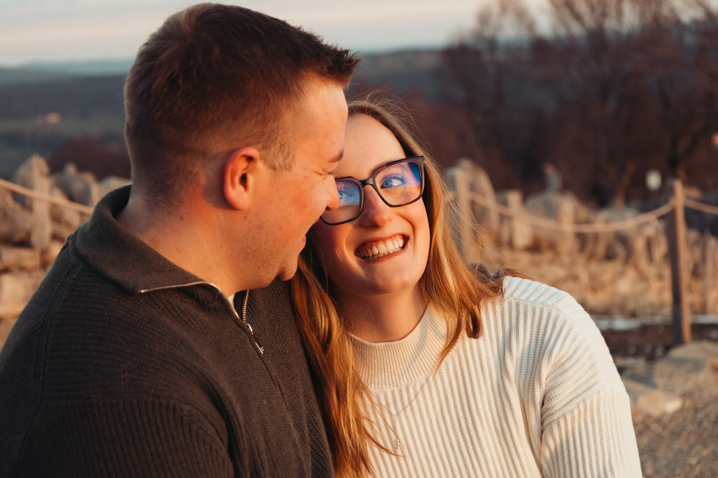 Un couple souriant, la femme portant des lunettes, se prend en photo ensemble lors d'une promenade en extérieur au crépuscule.