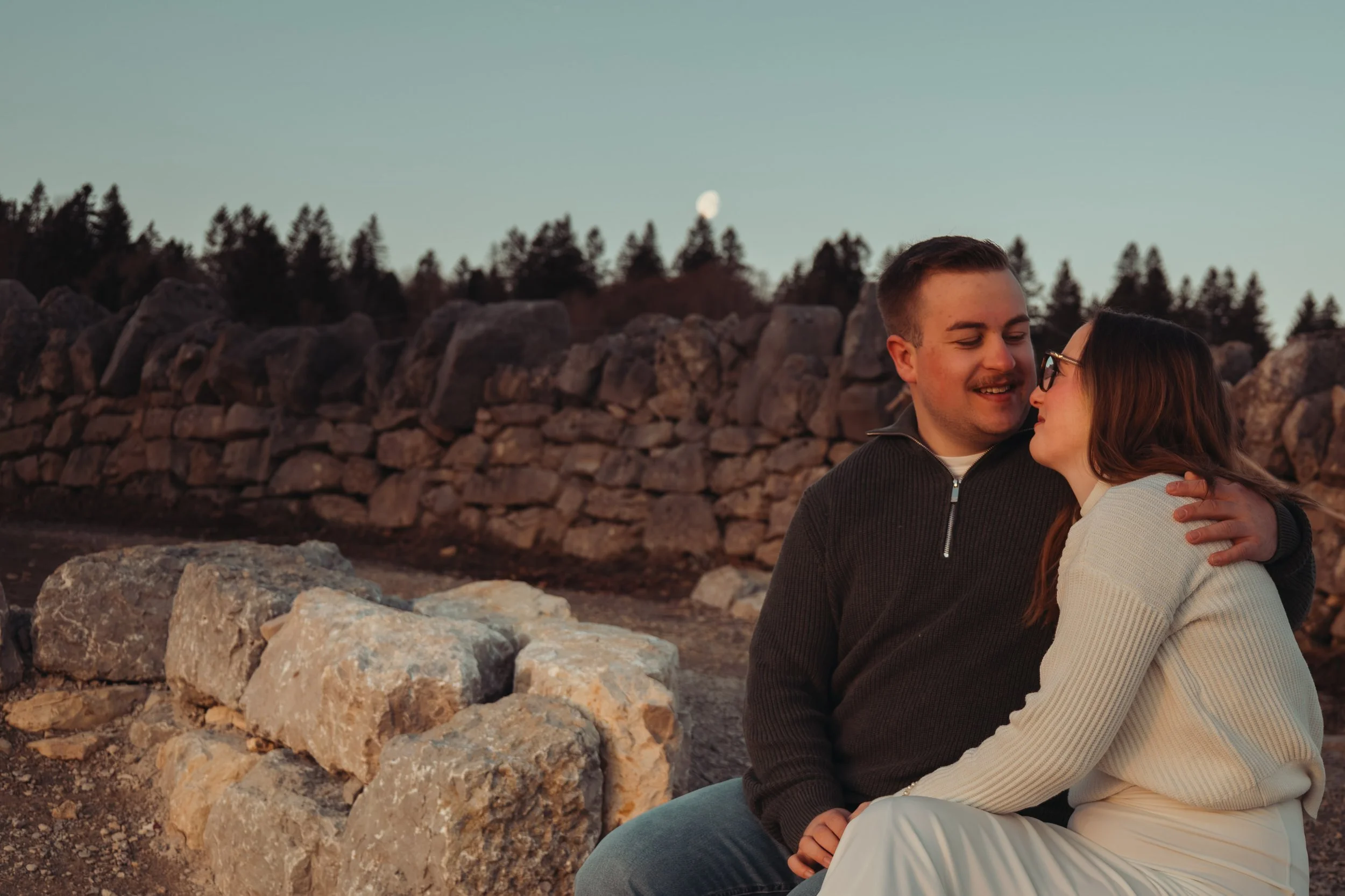 Un couple assis près d'une pile de pierres, amis ou en couple, partageant un moment intime en plein air avec un ciel clair et une lune visible en arrière-plan.