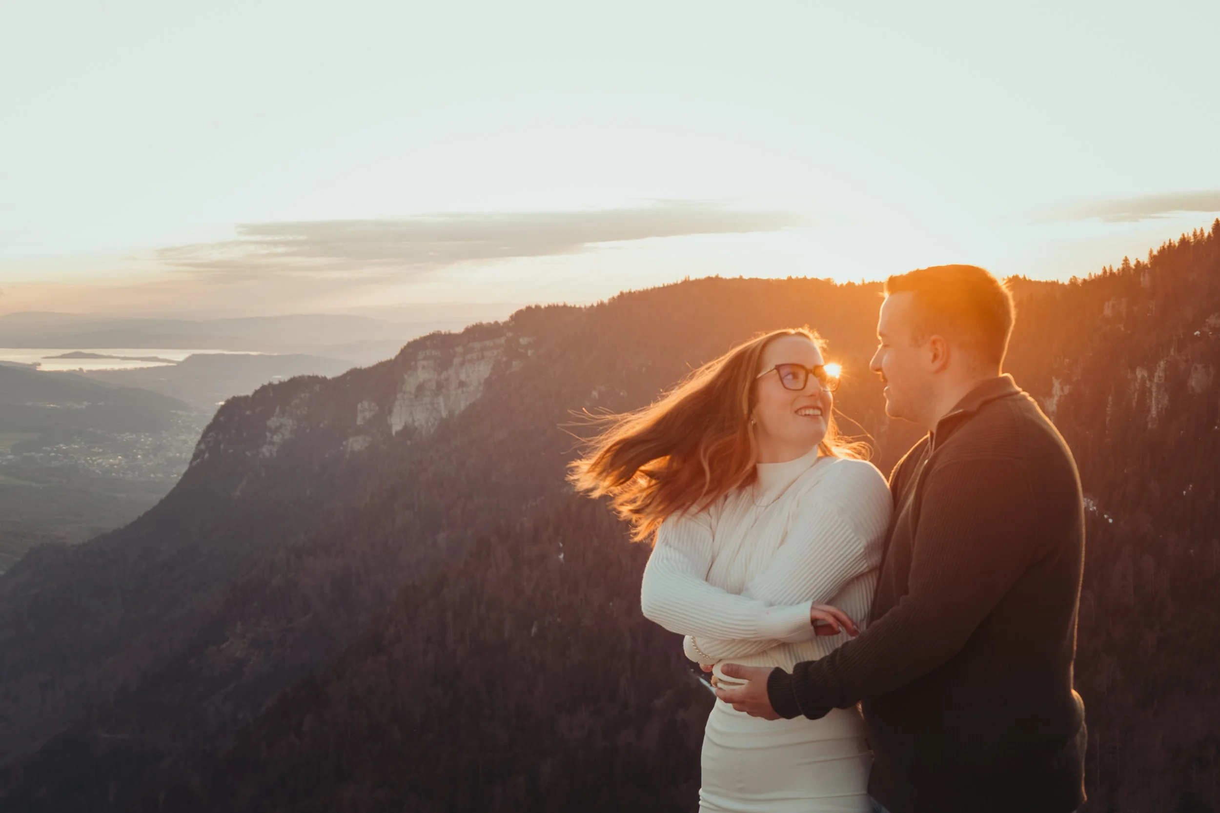 Un couple qui sourit, se tenant la main, avec une vue sur une vallée et des montagnes au coucher du soleil.