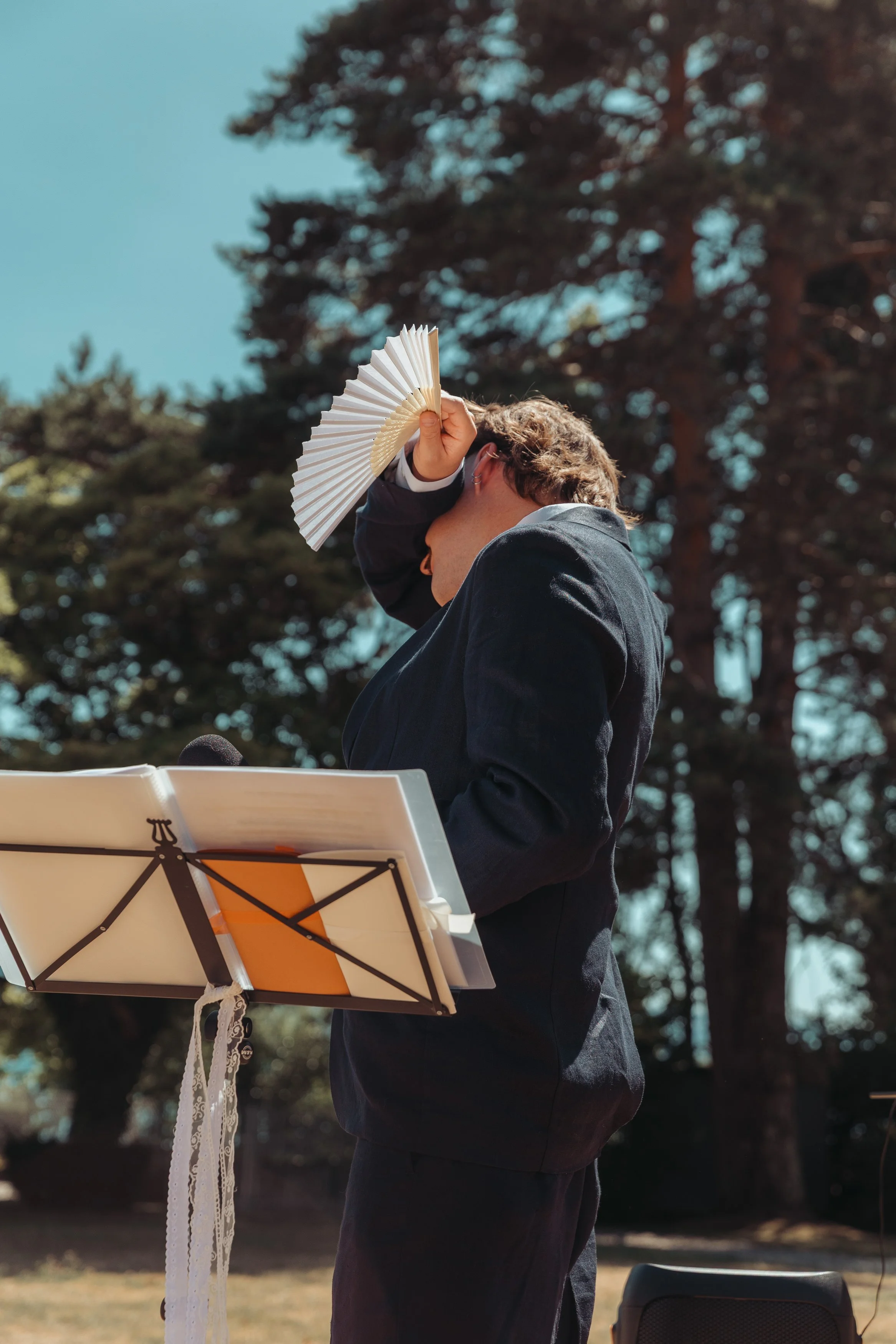 Une personne en costume noir joue du piano à l'extérieur, tenant une éventail blanc, avec un pupitre de musique devant elle et un arbre en arrière-plan.
