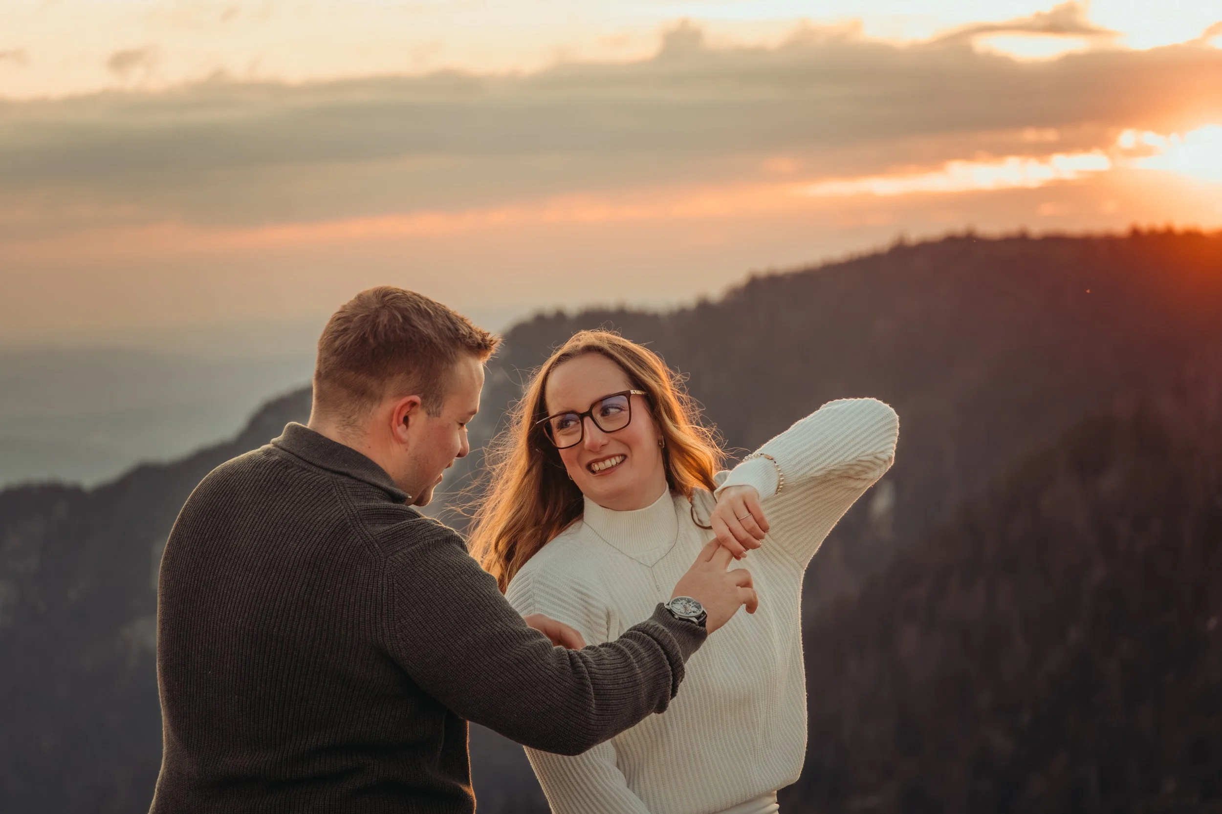 Un jeune couple riant et posant ensemble lors du coucher du soleil en plein air, avec des montagnes en arrière-plan.