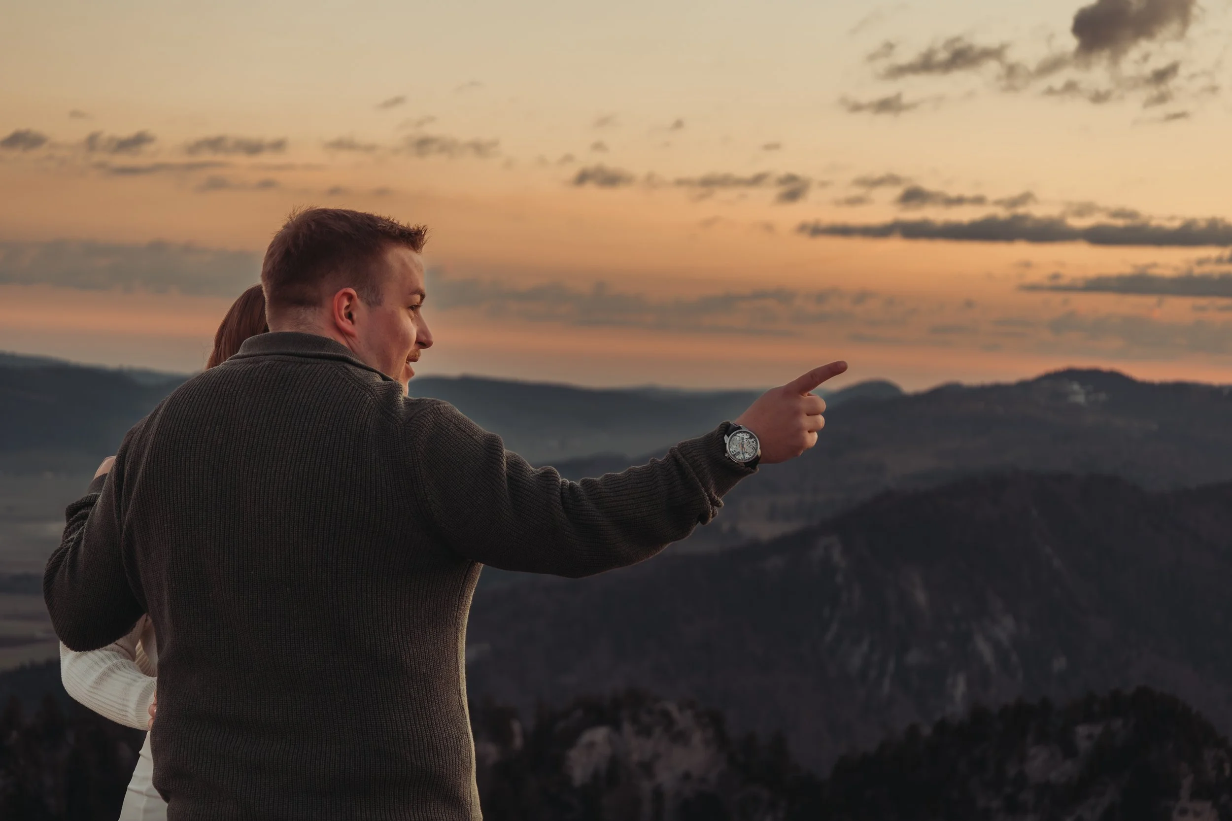 Deux personnes regardent un coucher de soleil sur une région montagneuse, l'homme porte une montre et pointe du doigt vers l'horizon.
