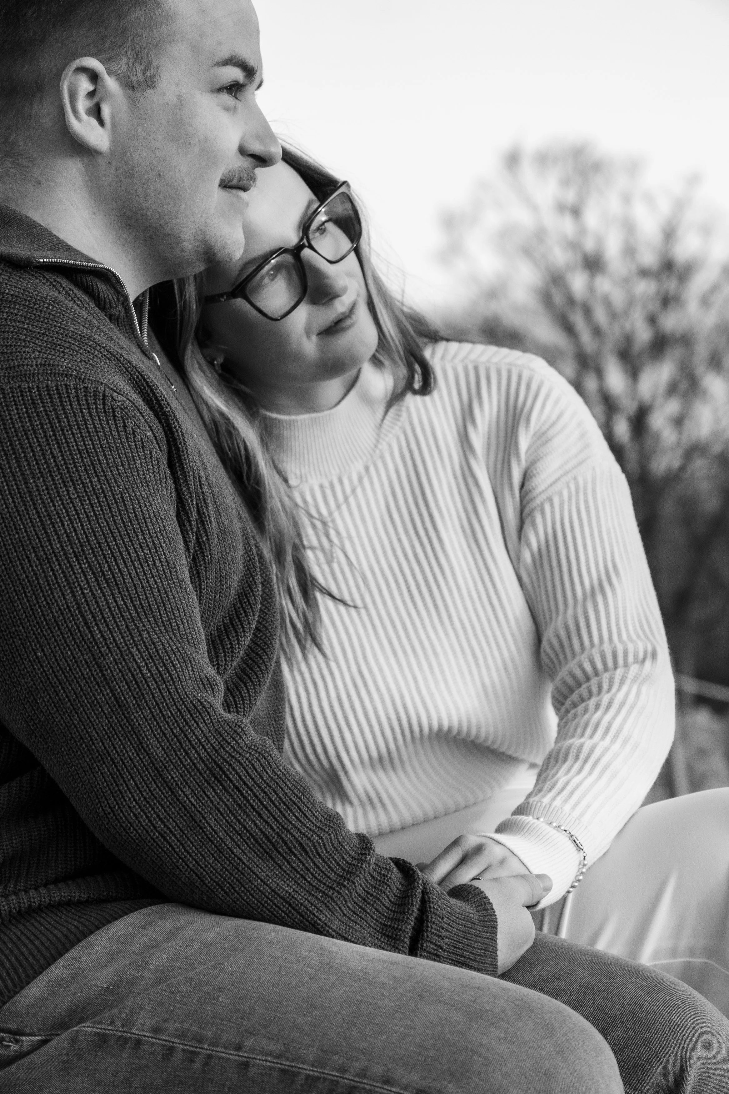 Deux personnes assises côte à côte, une femme et un homme, semblent partager un moment de proximité et de tendresse, avec l'homme touchant la main de la femme. L'image est en noir et blanc, avec un arbre flou en arrière-plan.