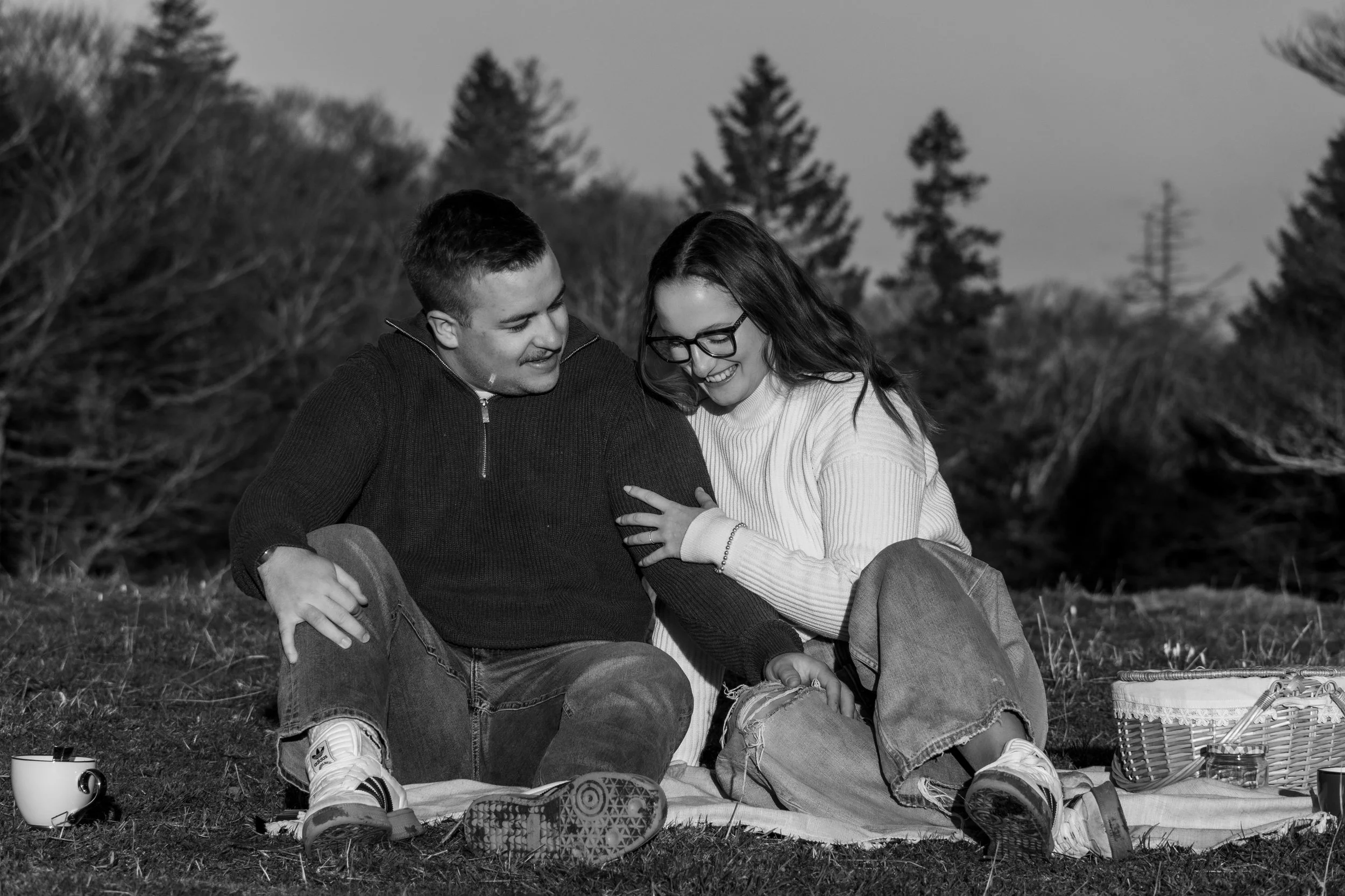 Deux jeunes adultes assis sur l'herbe dans un parc, riant et partageant un moment de complicité, avec des arbres en arrière-plan.