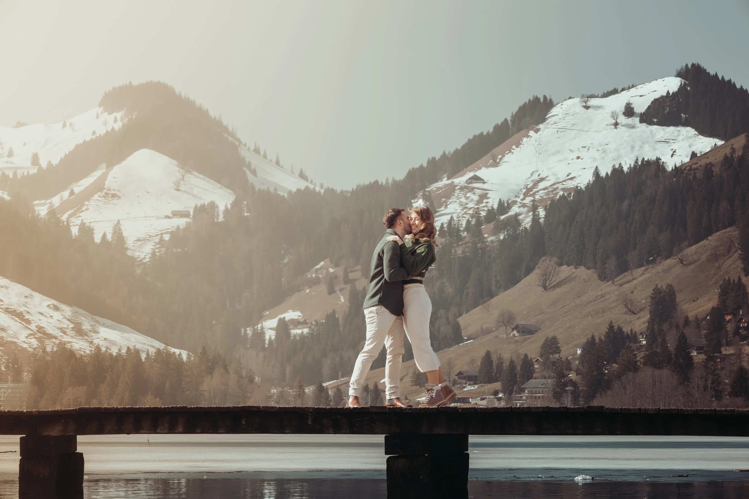 Un couple qui s'embrasse sur un pont en bois surplombant une rivière dans un environnement montagneux enneigé avec des forêts.