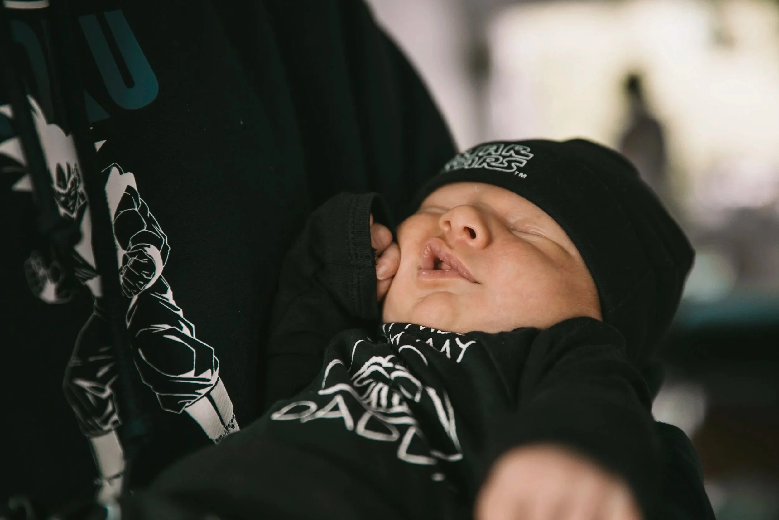 Bébé endormi dans un porte-bébé, portant un bonnet noir et des vêtements noirs avec un motif Star Wars.