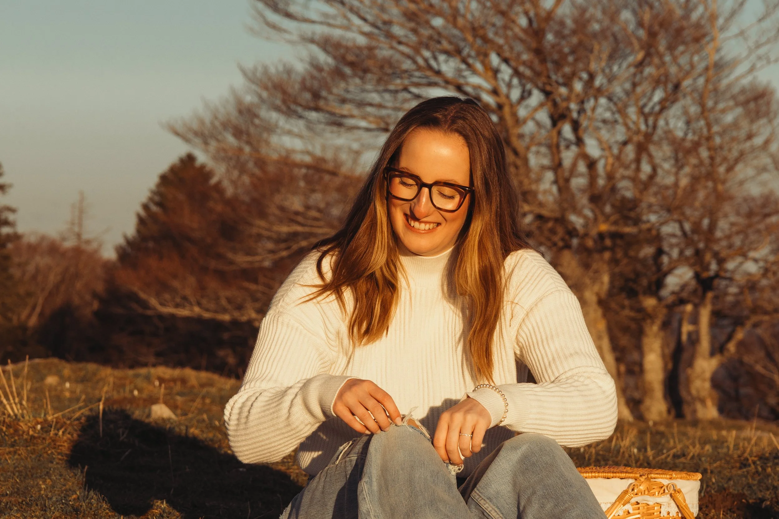 Une jeune femme souriante, portant des lunettes, assise dehors en automne, avec des arbres sans feuilles en arrière-plan.