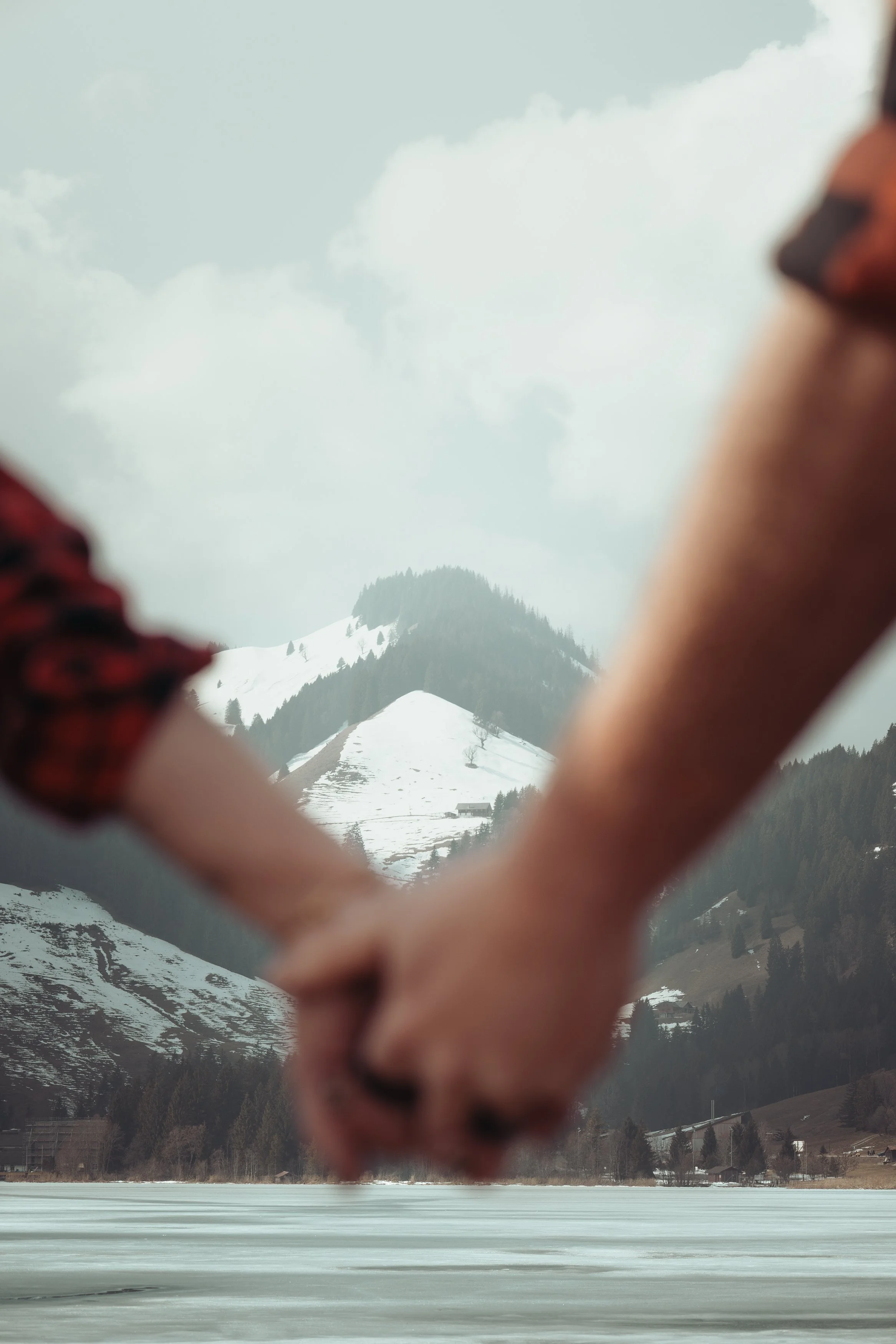 Deux personnes qui tiennent la main dans un paysage de montagnes enneigées et un lac gelé, avec des nuages dans le ciel.