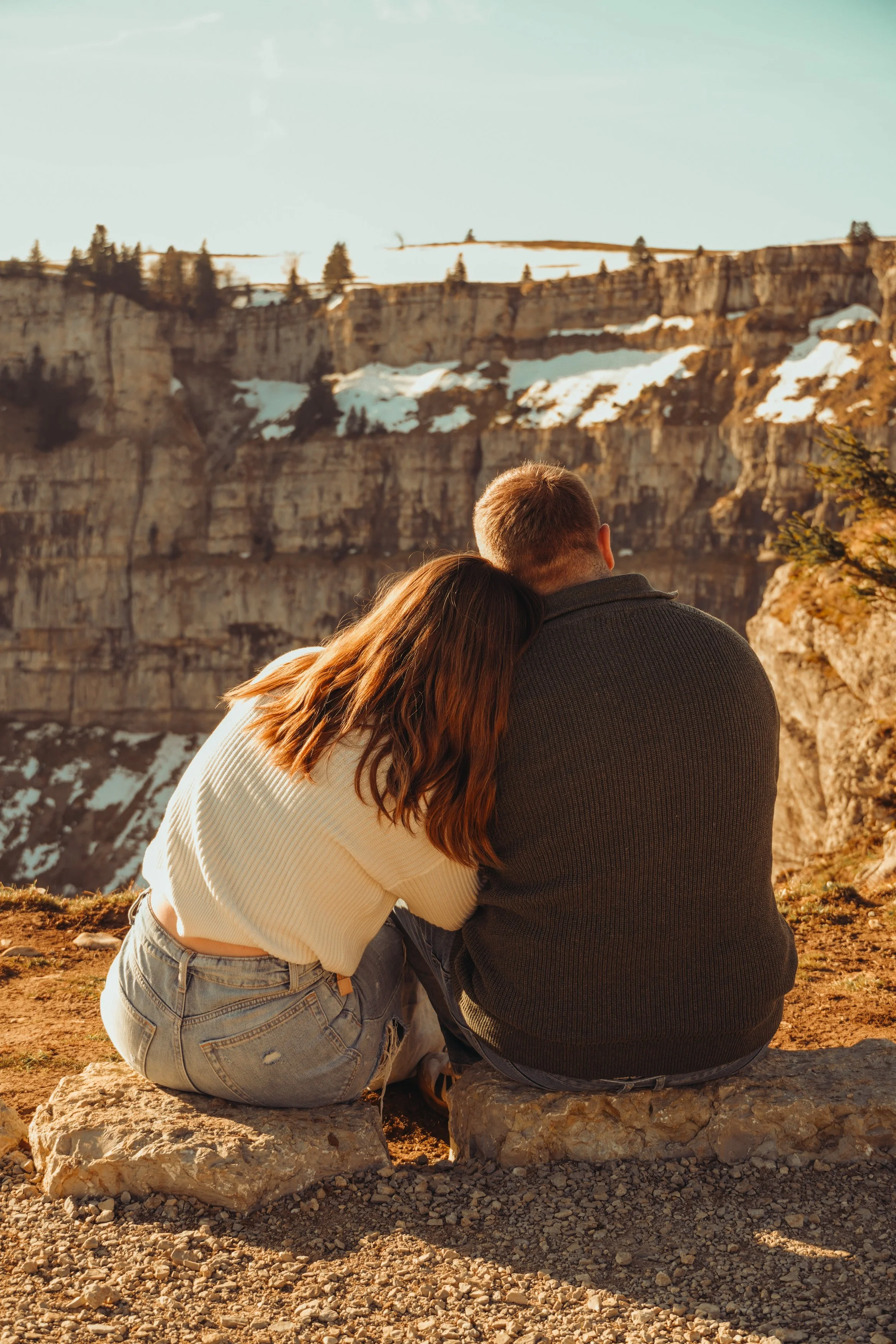Un couple assis sur un rocher, regardant un paysage de falaises avec de la neige, sous un ciel clair.