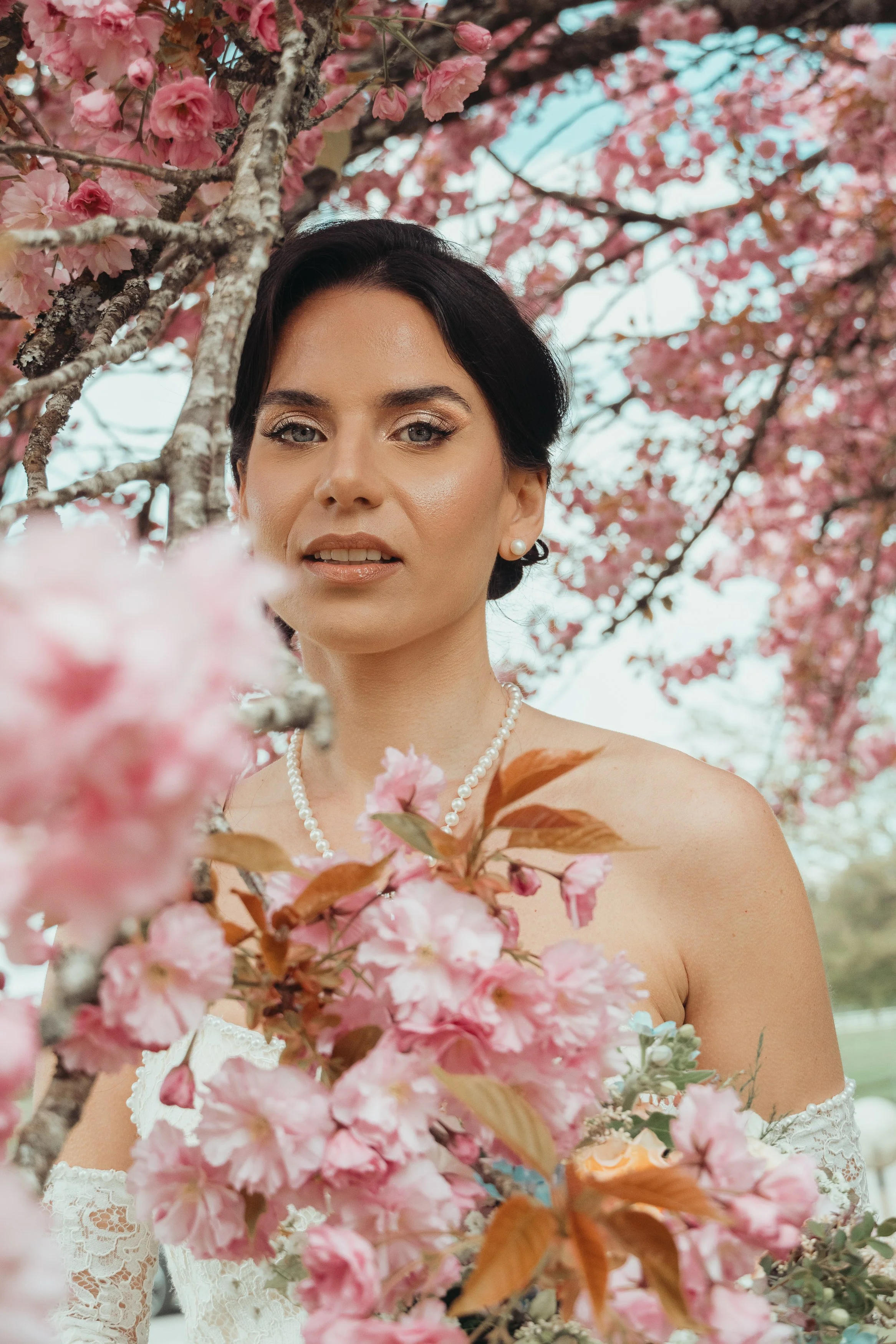 Une femme portant un collier de perles et une robe blanche en dentelle, se tenant devant un arbre en fleurs roses.
