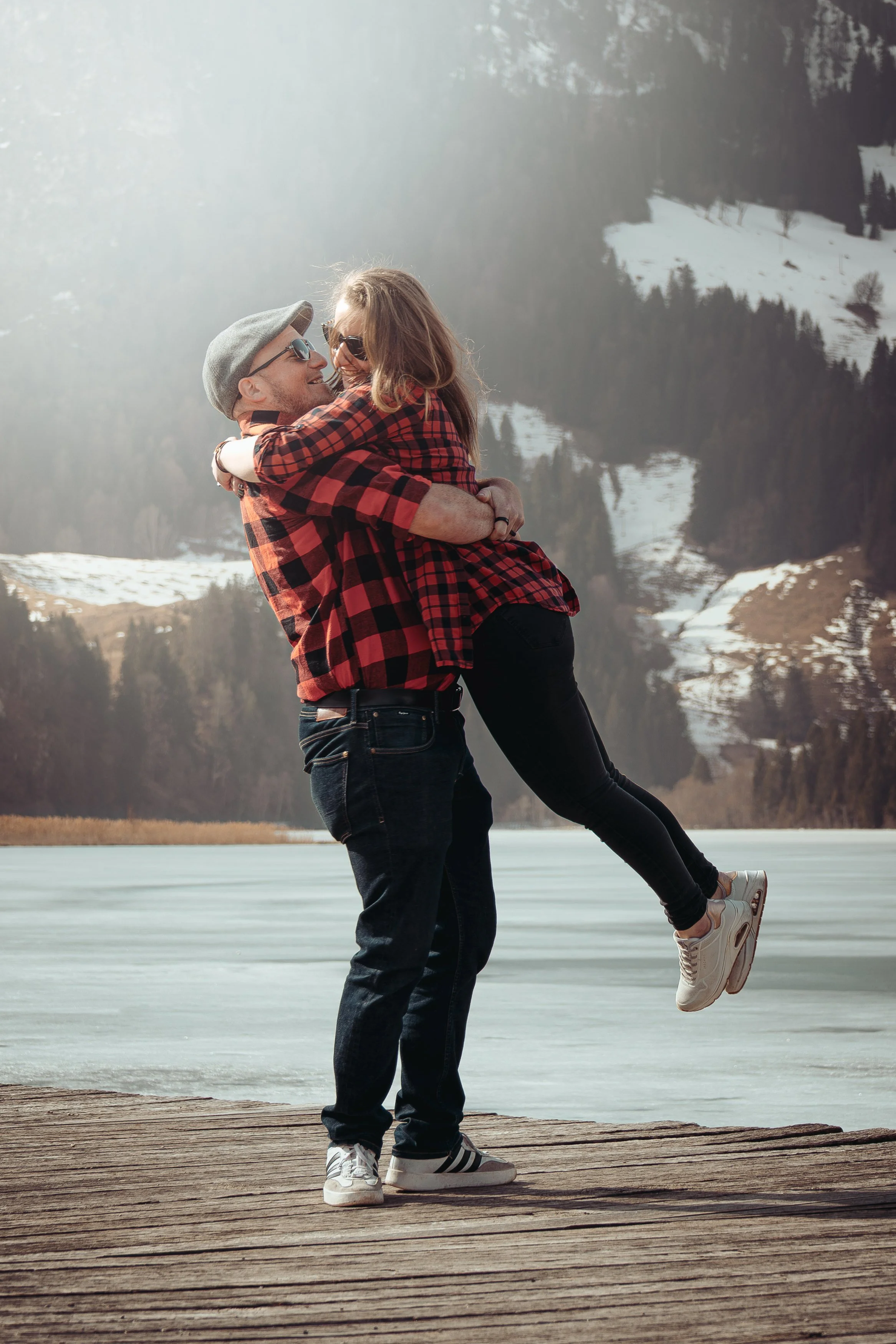 Un couple en vêtements à carreaux, le homme tenant la femme dans ses bras devant un lac gelé et des montagnes enneigées.