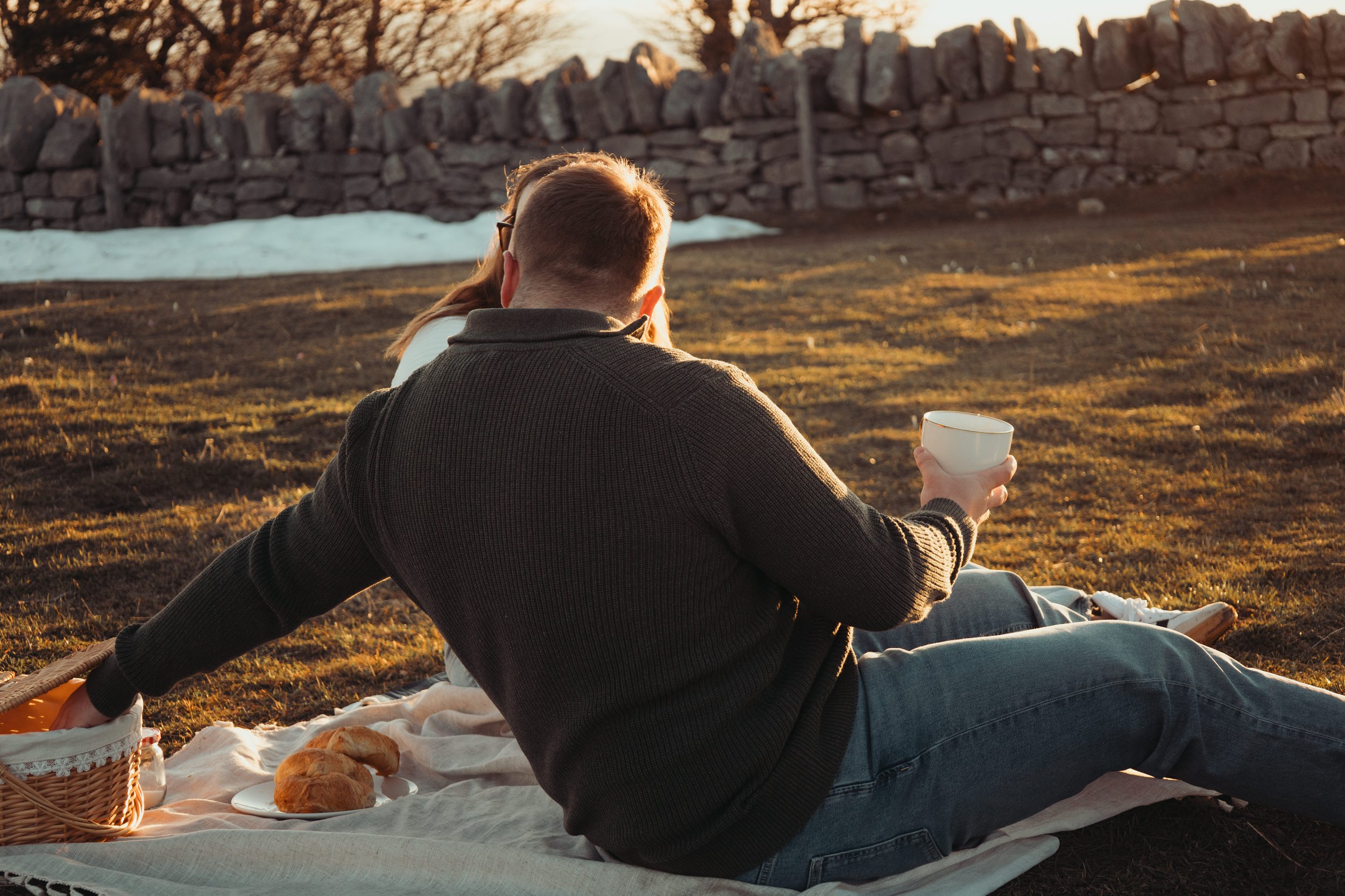 Deux personnes sont assises sur une couverture lors d'un pique-nique au coucher du soleil dans un champ, avec un panier, du pain et une tasse.