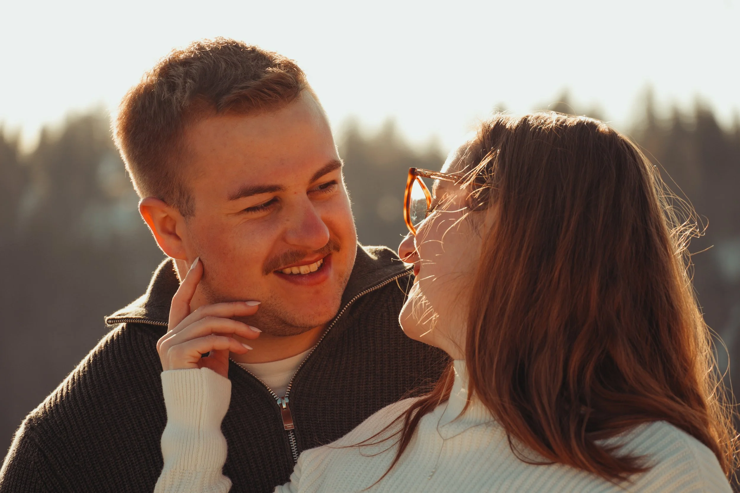 Un couple souriant se regarde tendrement, à l'extérieur, en plein jour, avec un arrière-plan flou de nature ou forêt.