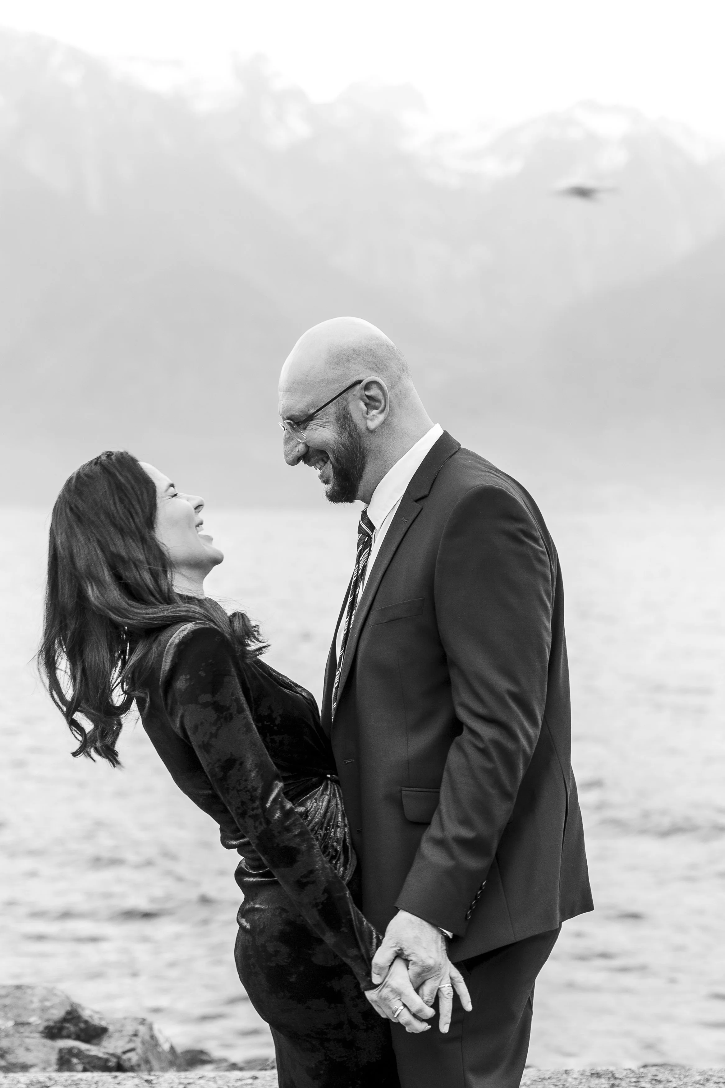 Un couple en costume, un homme et une femme, se regardant avec amour au bord d'un lac, en arrière-plan des montagnes, en noir et blanc.