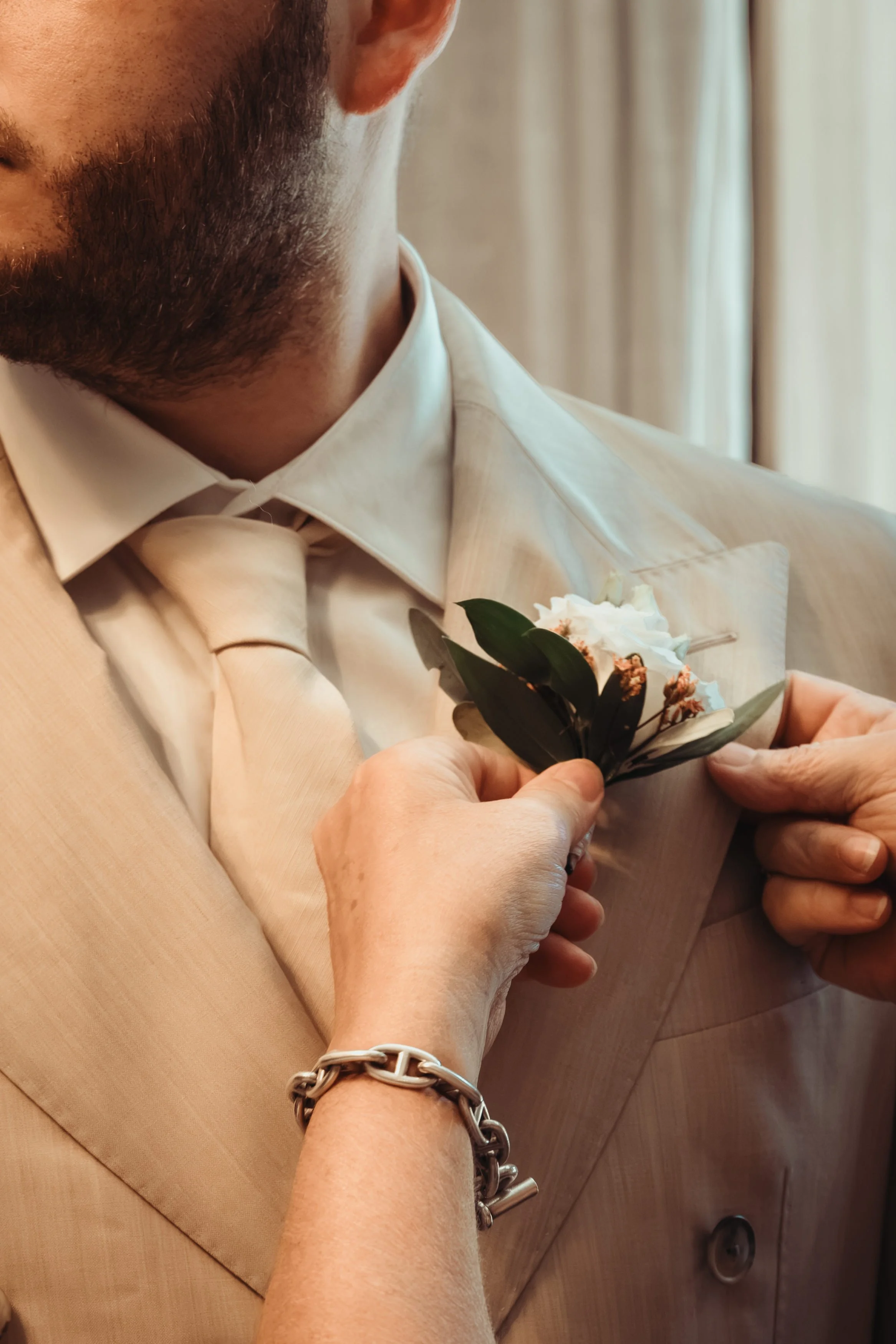 Un homme en costume beige tenant une petite bouquet de fleurs blanc avec des feuilles vertes.