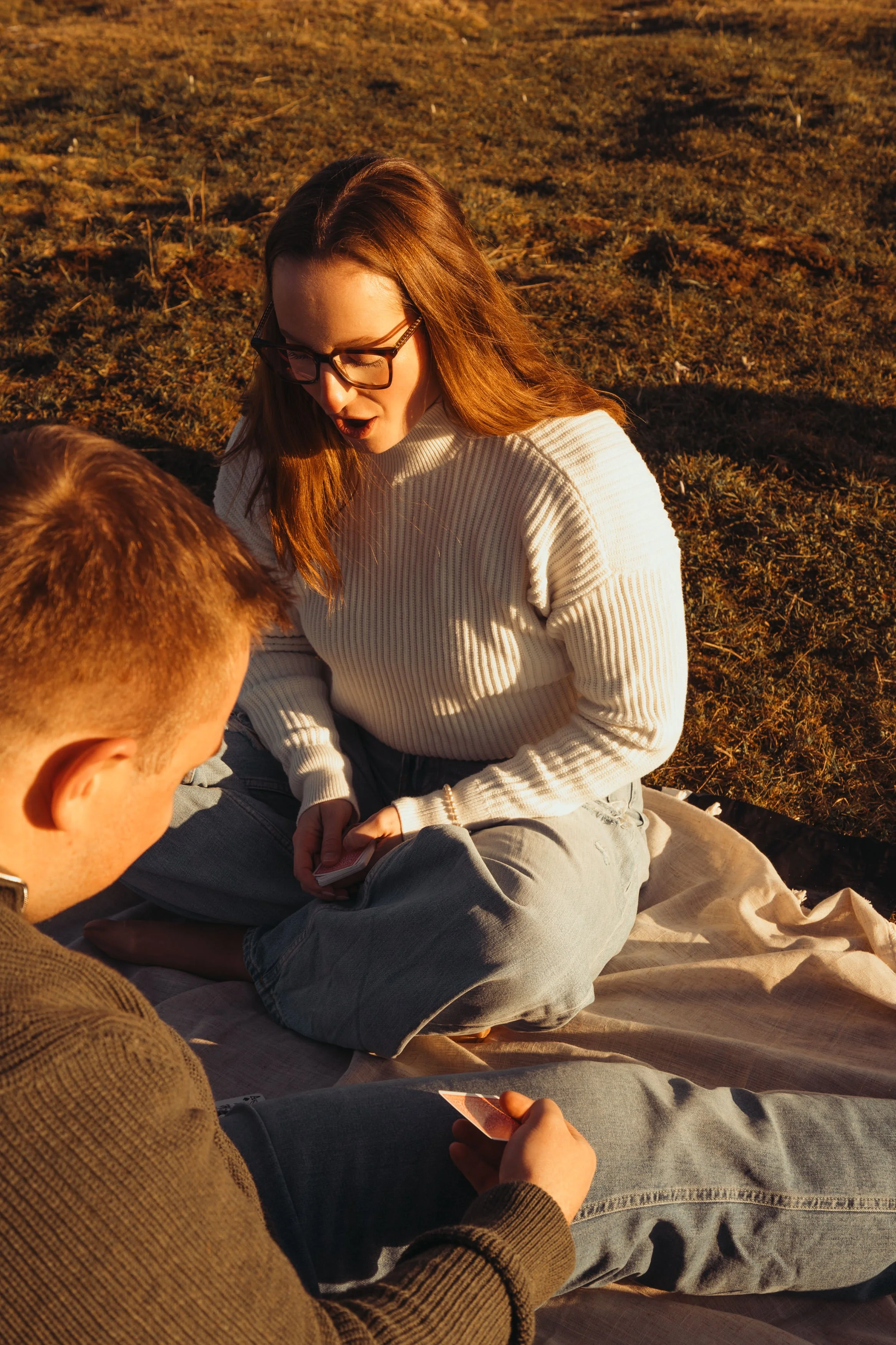 Deux jeunes adultes assis sur une couverture dans un champ, personne à droite avec des cheveux roux et lunettes, portant un pull blanc, personne à gauche avec des cheveux courts, portant un pull marron, discutant ou jouant à un jeu de cartes lors du 