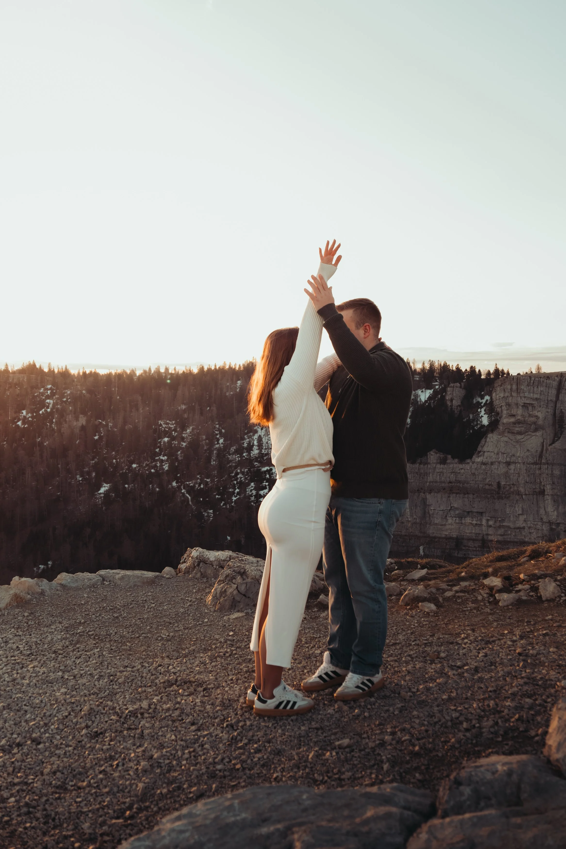 Un couple debout près d'une falaise, s'embrassant lors d'un coucher de soleil dans un paysage rocheux avec des montagnes en arrière-plan.