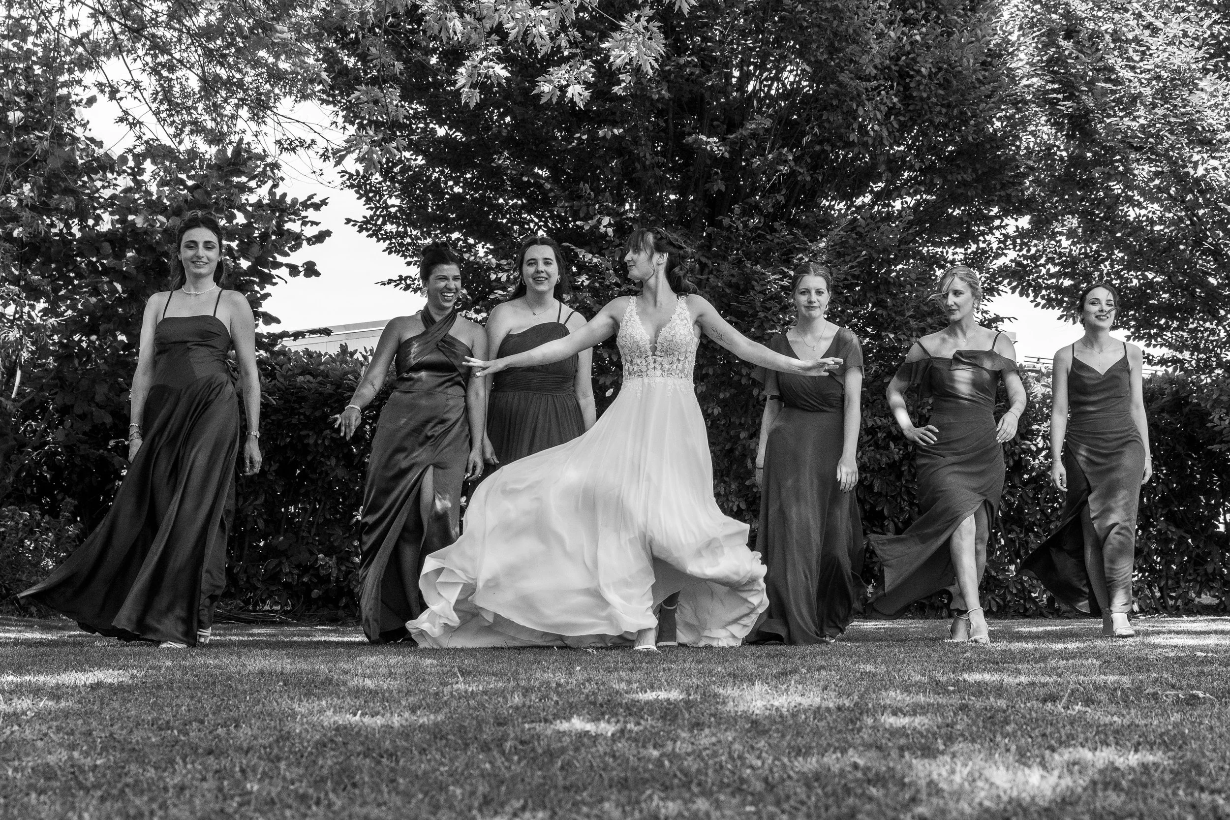 Un groupe de femmes habillées en robes de soirée, probablement lors d'un mariage, marchant ou posant dans un jardin ombragé avec des arbres en arrière-plan.