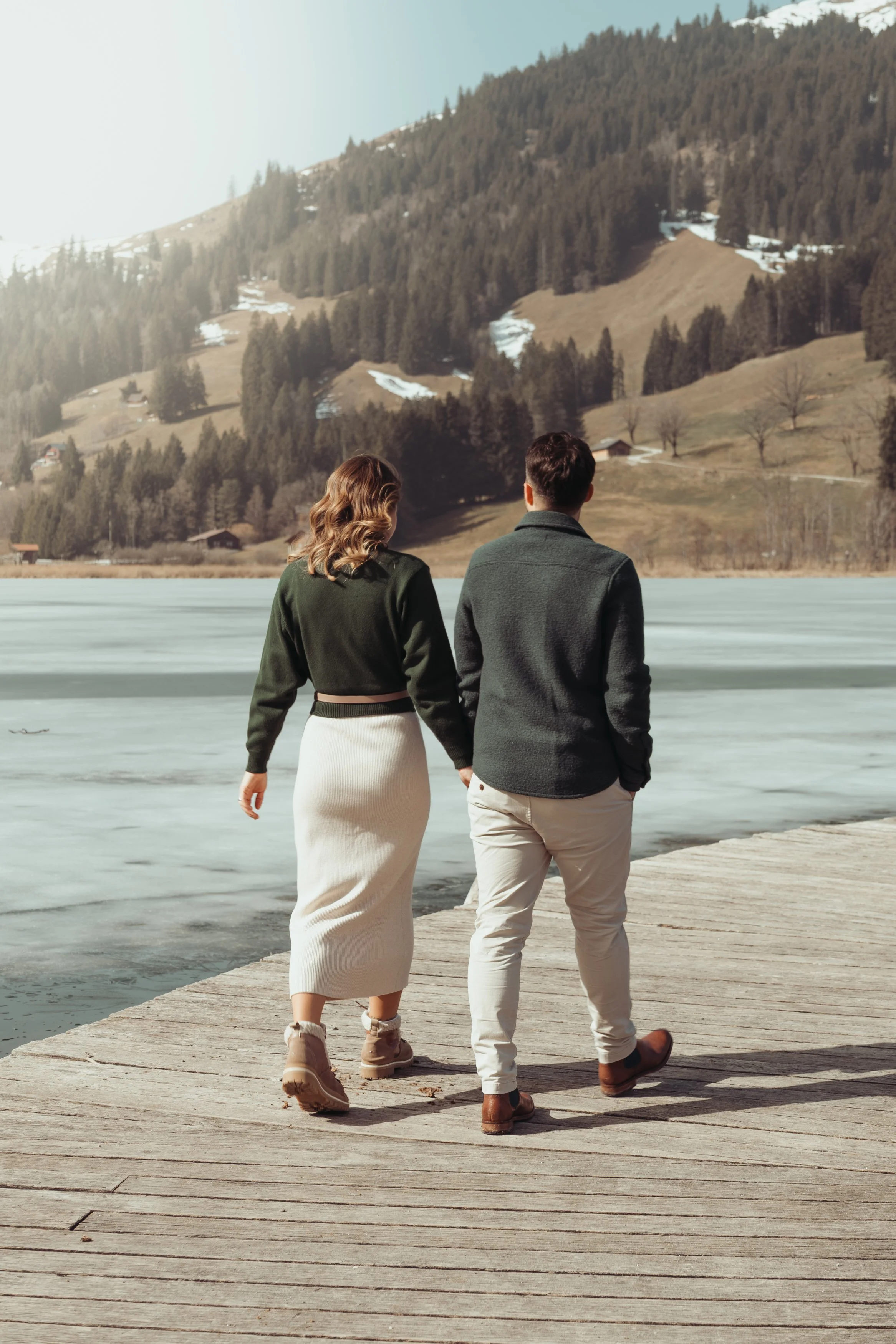Un couple marche sur un pont en bois au bord d'un lac, avec des montagnes et des arbres en arrière-plan.
