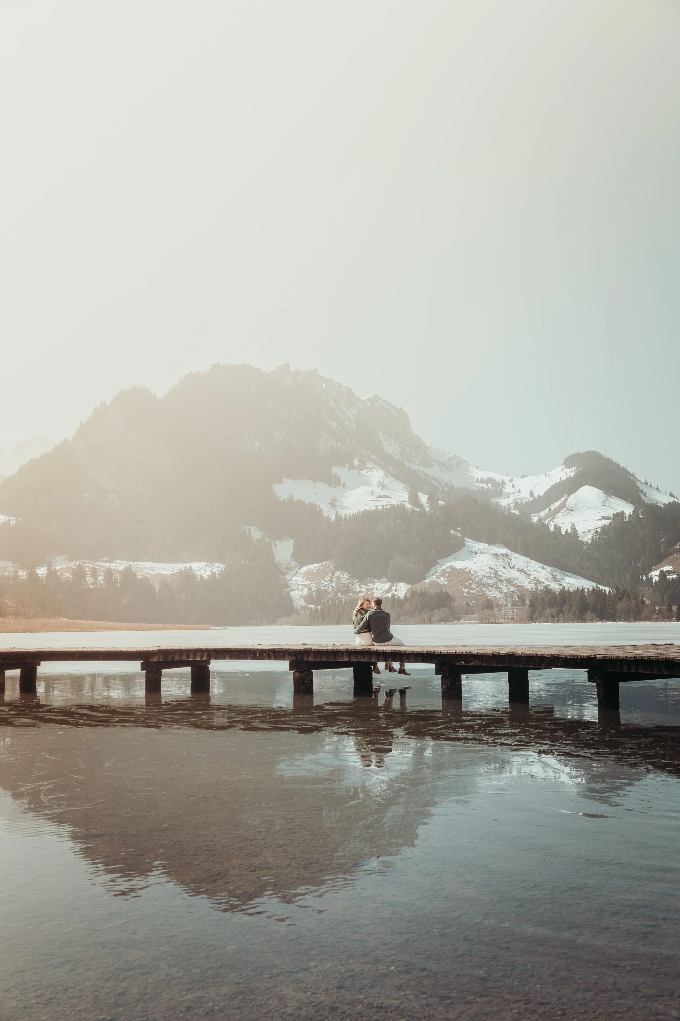 Deux personnes assises sur une jetée en bois, entourées de montagnes enneigées et d'un lac calme, avec un ciel clair.