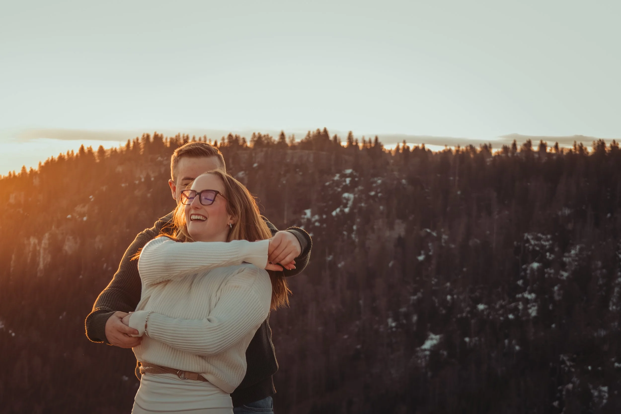 Un couple partage un moment d'affection en plein air, avec arrière-plan de montagnes enneigées et d'arbres lors du coucher de soleil.