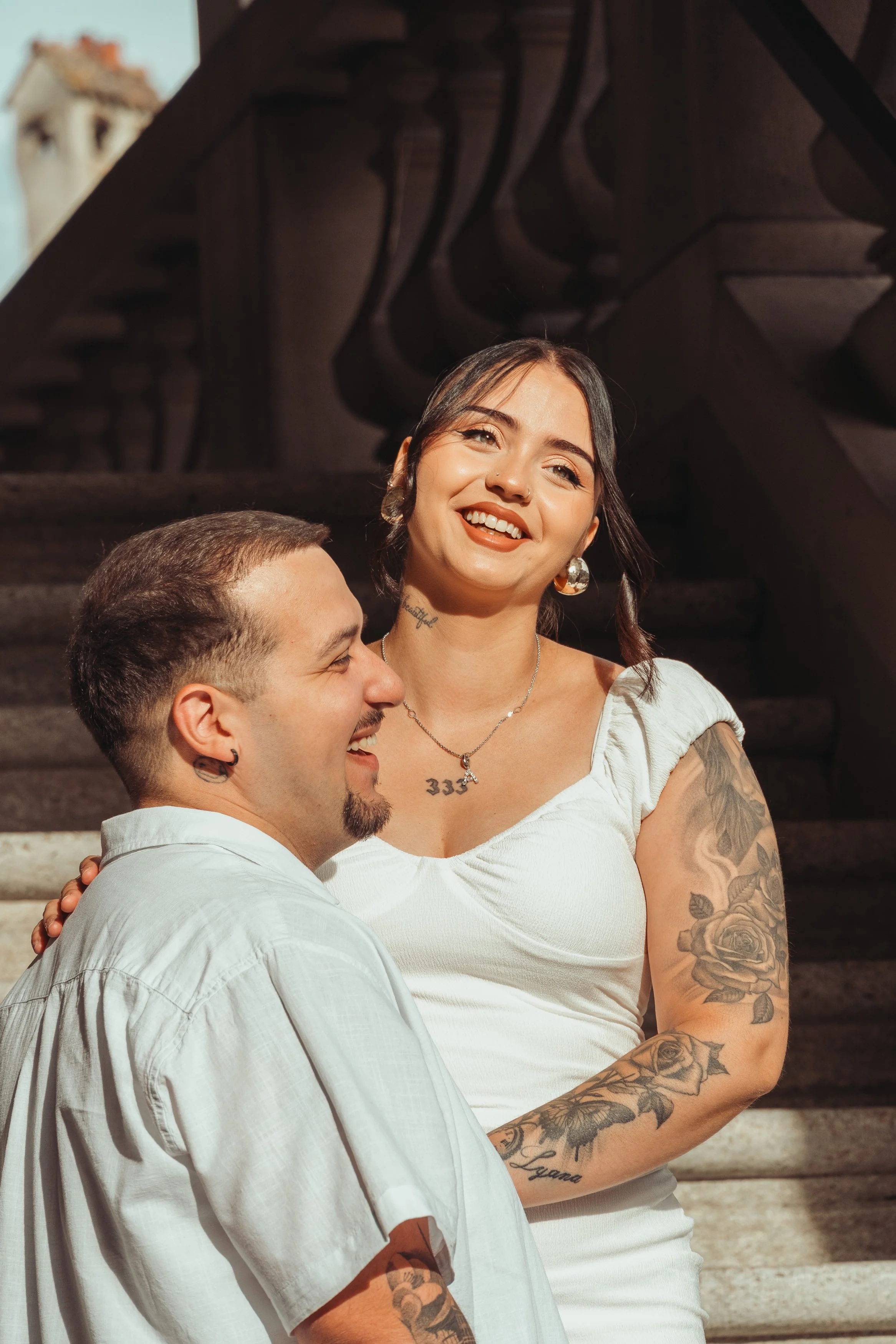 Un couple souriant, la femme en white t-shirt avec tatouages sur le bras, le homme en chemise blanche, devant un escalier en bois.